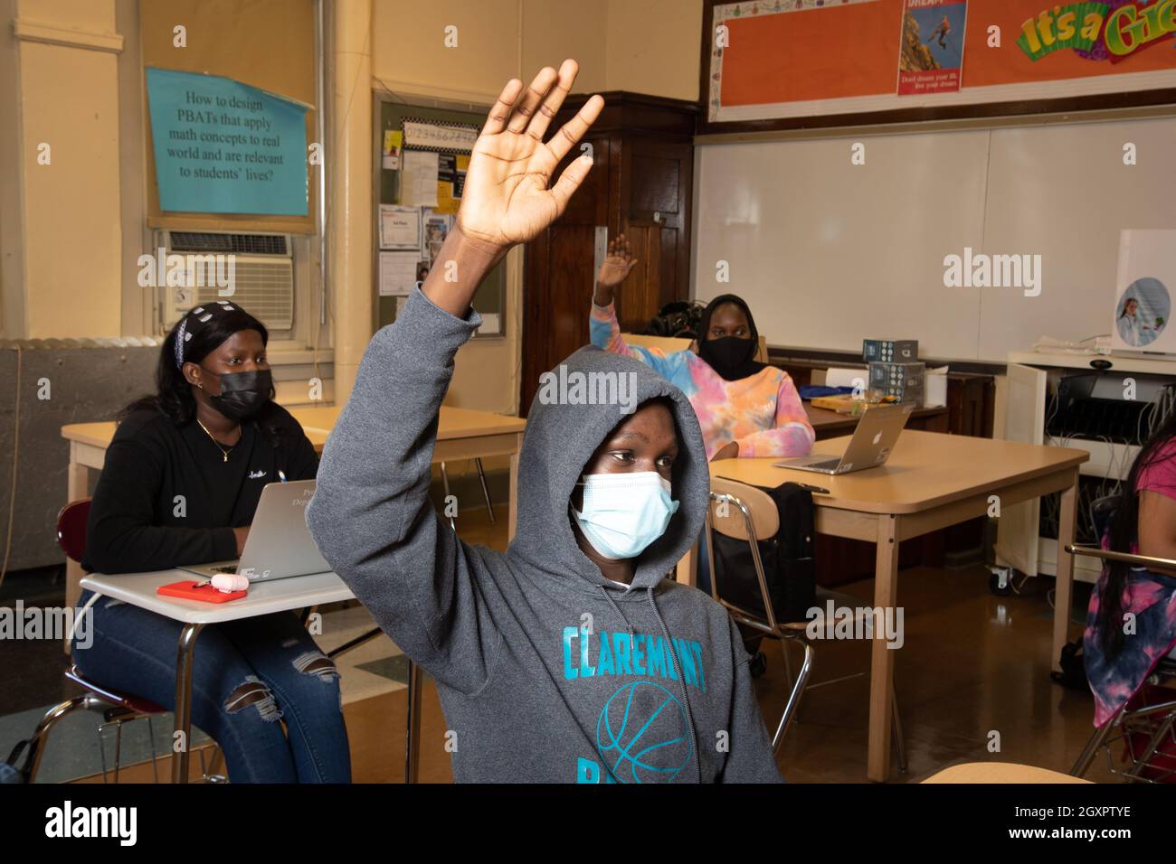 Istruzione High School scena in aula, studente maschio alzando la mano per rispondere alla domanda, studenti femmina dietro di lui, uno alzando la mano maschere facciali Covid Foto Stock