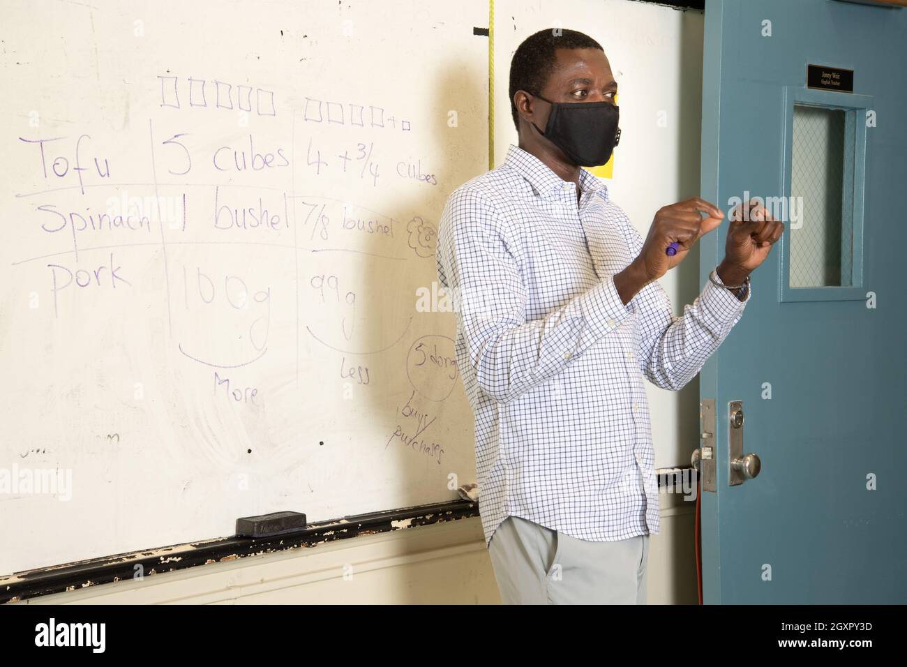 Istruzione High School scena in aula, insegnante maschile che presenta, in piedi di fronte a bordo di cancellazione a secco, gesturando come lui parla, indossare maschera facciale Foto Stock