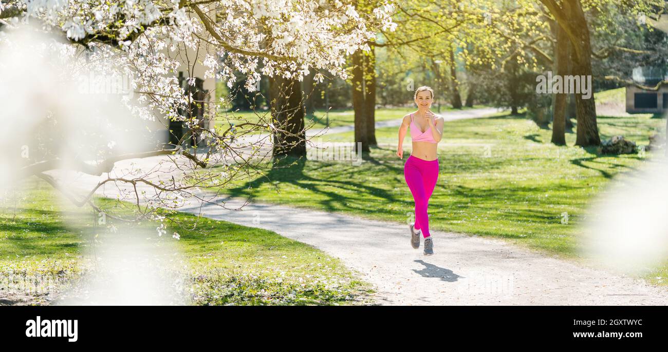 Fit donna che corre lungo un percorso verso la fotocamera durante la primavera visto attraverso la fioritura Foto Stock