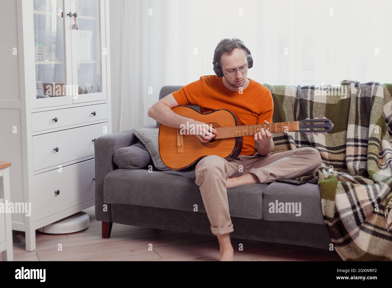 Giovane uomo caucasico in cuffia che suona la chitarra seduto sul divano a casa. Pratica la musica, stile di vita foto Foto Stock