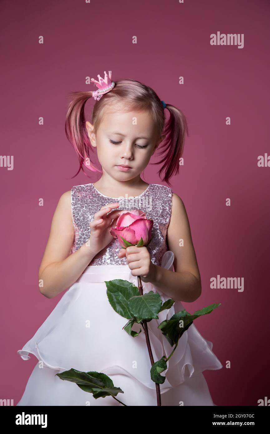 Piccola ragazza caucasica in un abito festivo con paillettes in posa con fiore di rosa su sfondo rosa guardando la fotocamera Foto Stock