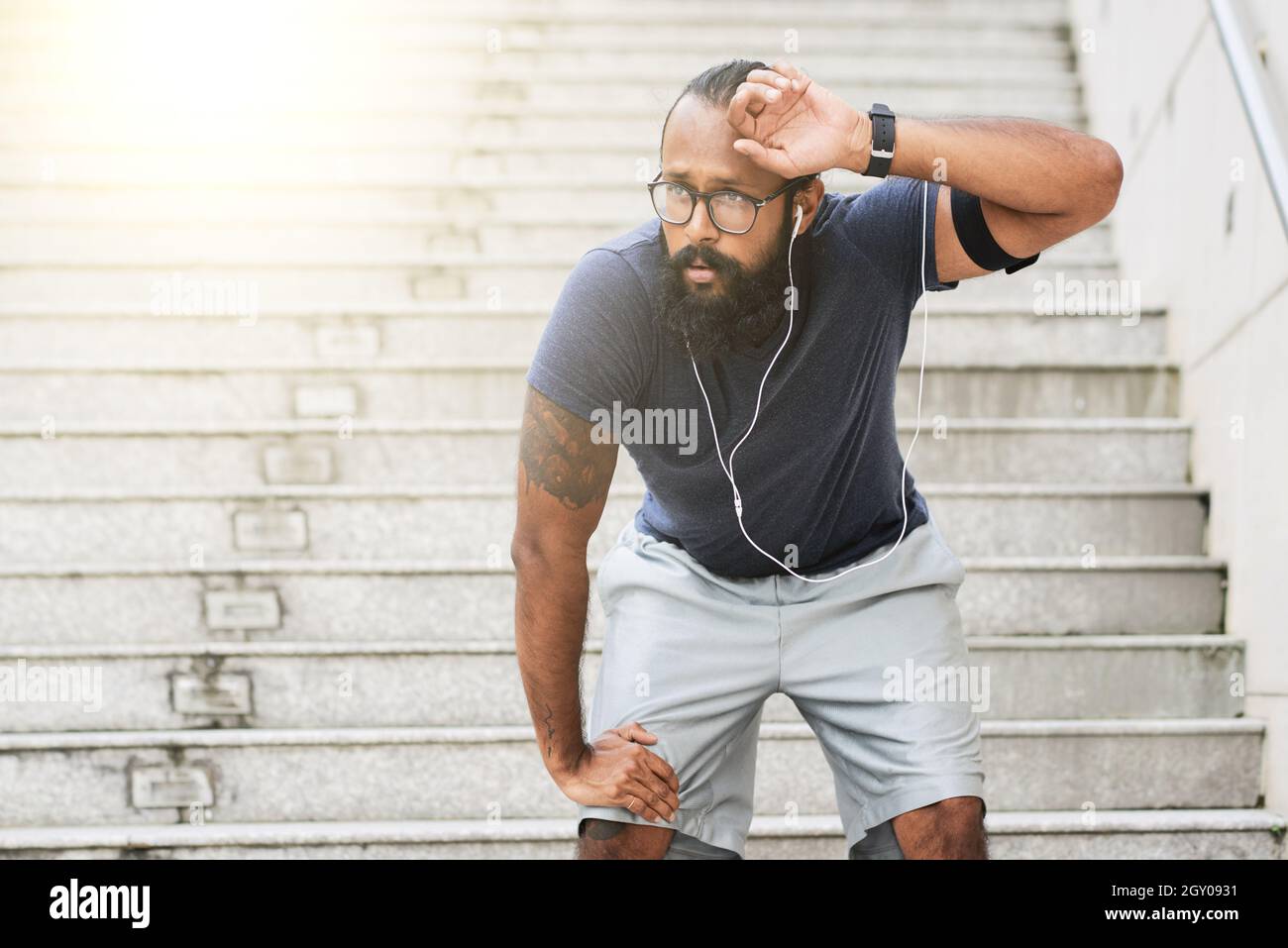 Giovane atleta indiano stanco in occhiali e cuffie in piedi contro le scale e sudore strofinare dopo la corsa Foto Stock