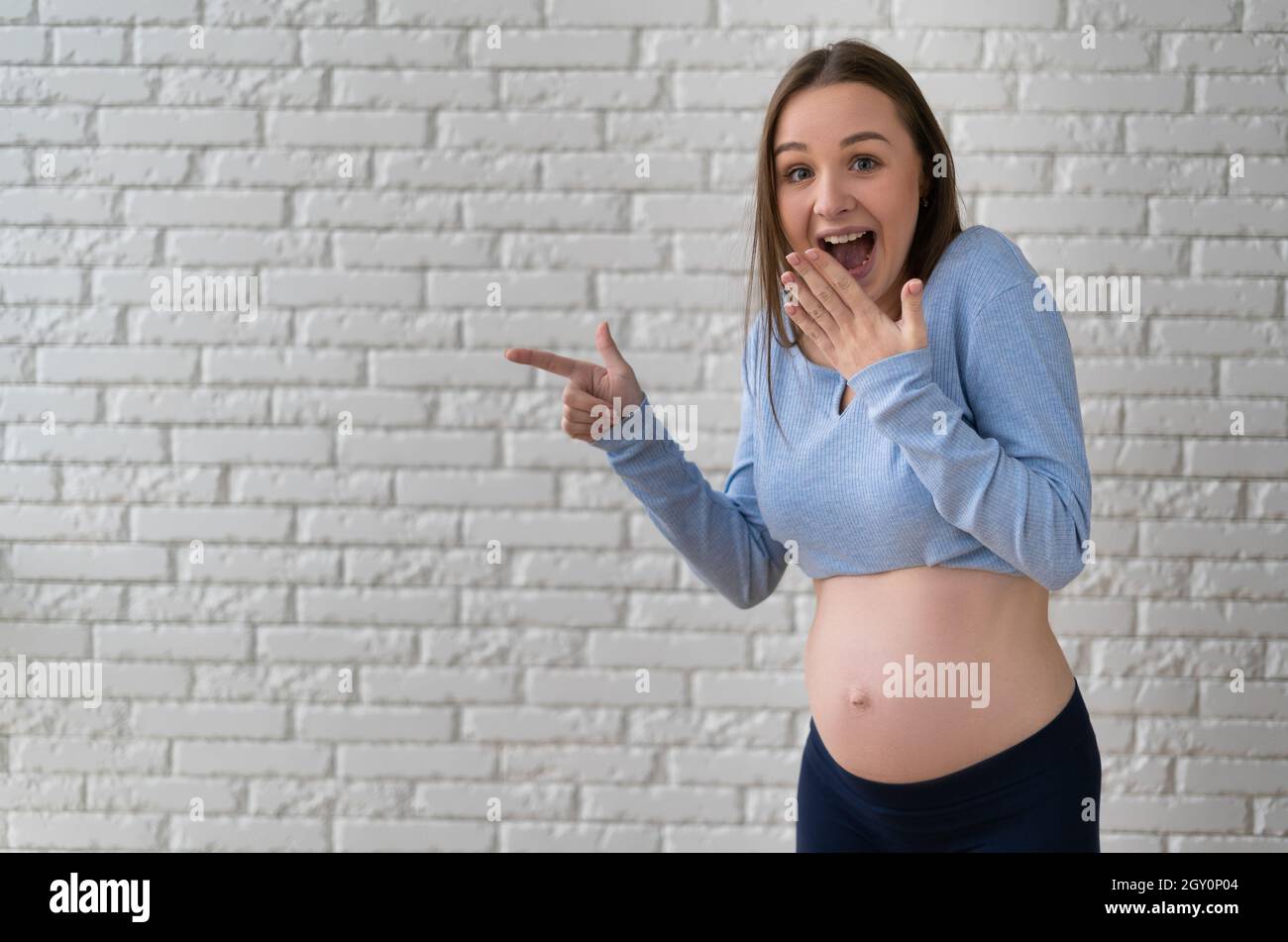 Ritratto di una giovane donna incinta. Si trova vicino ad un muro di mattoni, puntando il dito indice verso un punto vuoto. Foto Stock