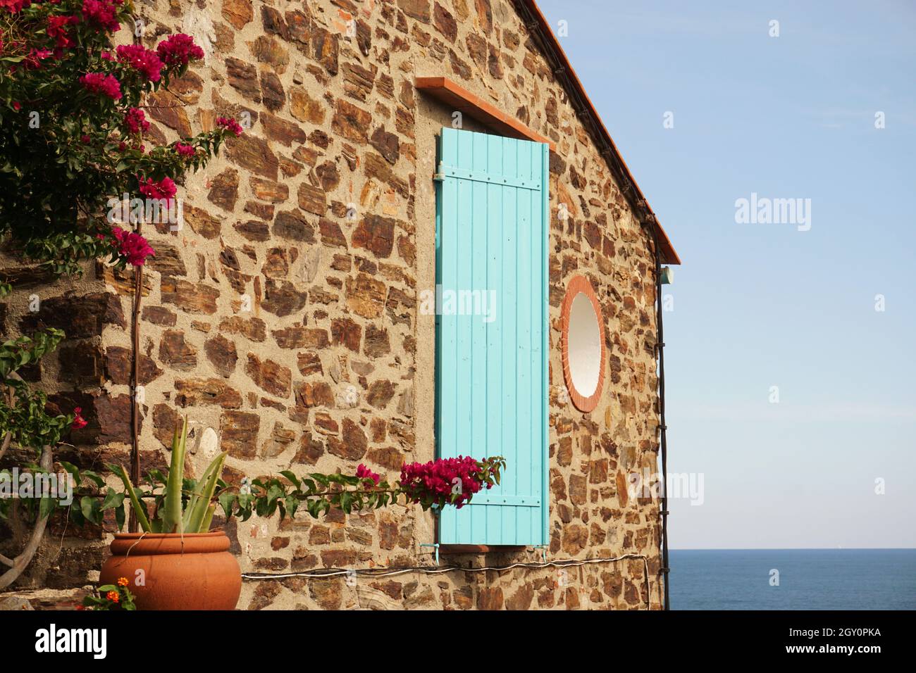 Parete a timpano di casa in pietra caratterizzata da persiane in legno color turchese e fiori rossi con mare Mediterraneo visibile. Collioure, Francia Foto Stock