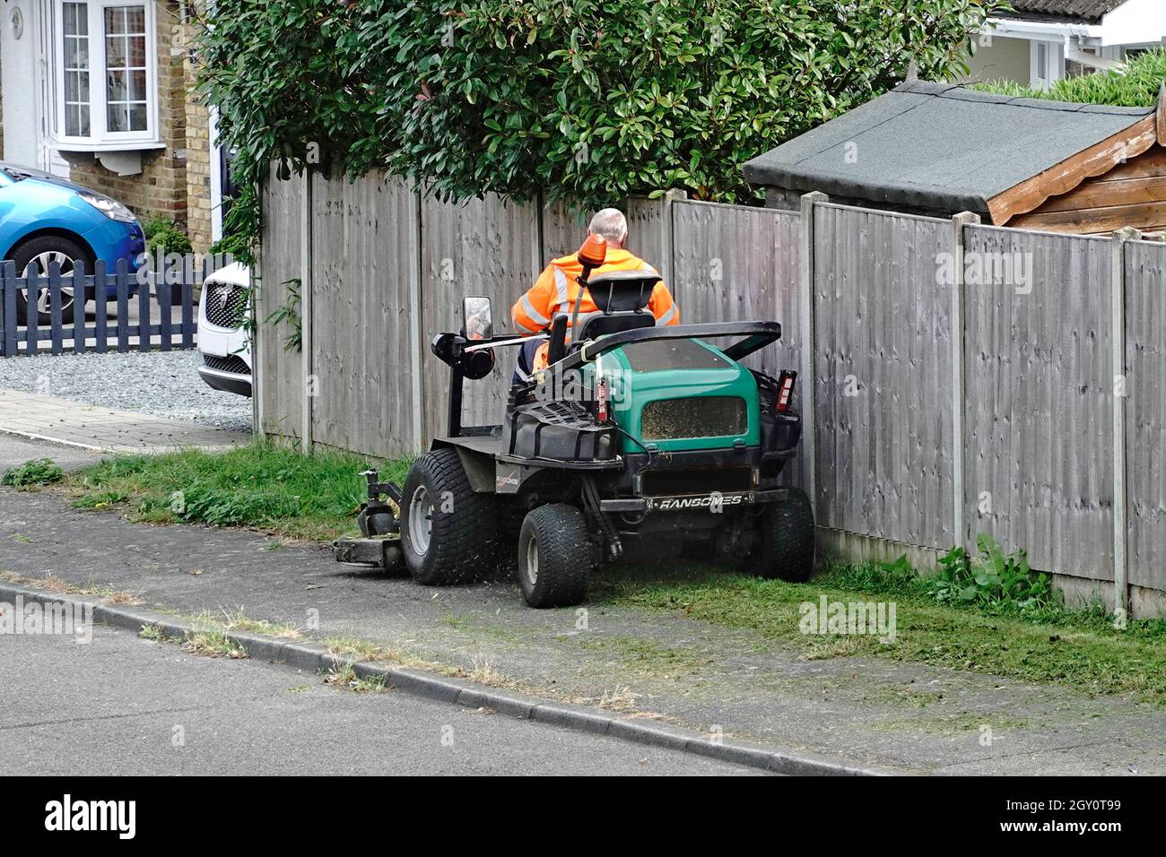 Uomo al lavoro che indossa giacca ad alta visibilità guidando una Ransomes quattro ruote si siede su erba falciatrice taglio council posseduto strada verge Essex Inghilterra UK Foto Stock