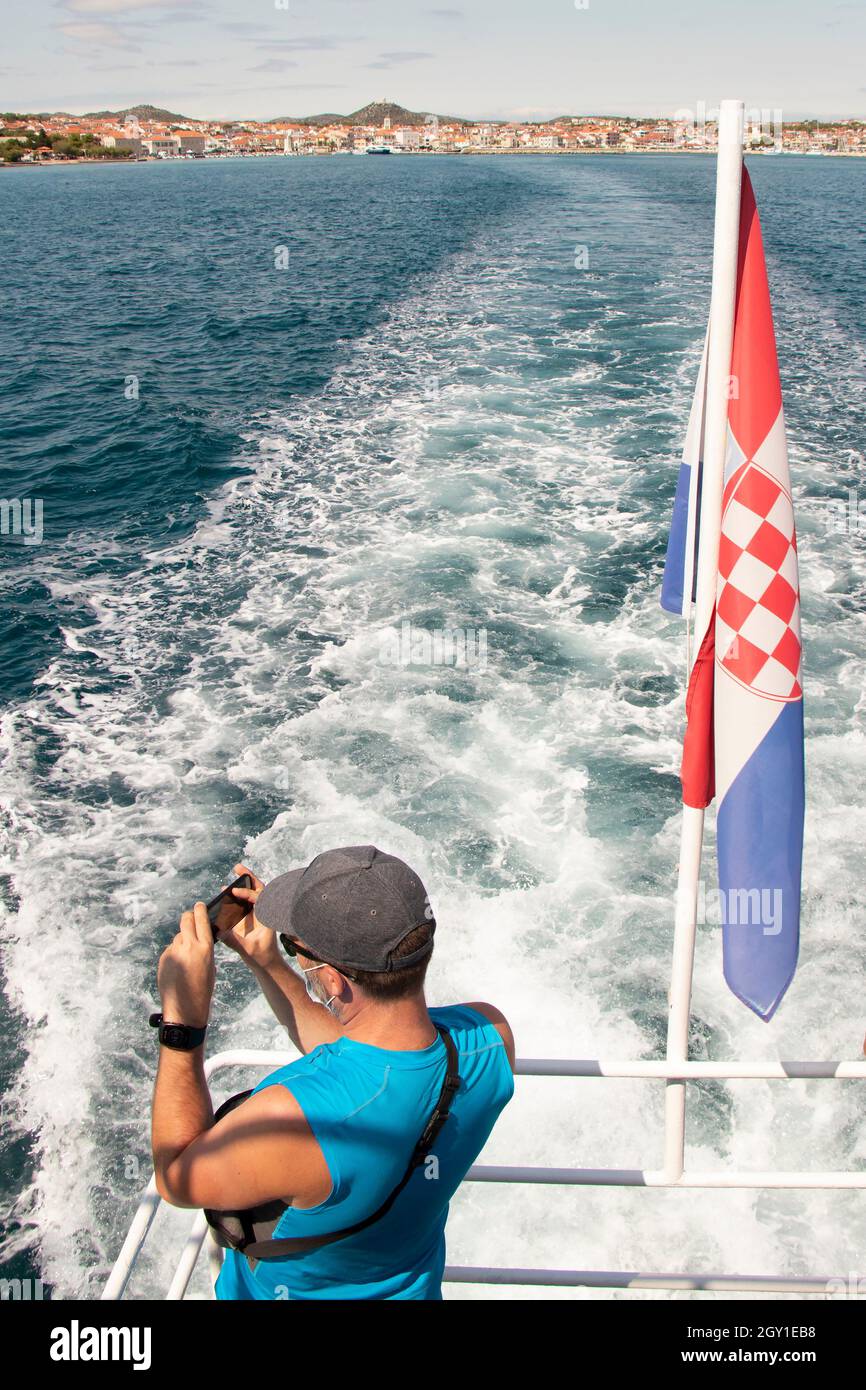 Vodice, Croazia - 25 agosto 2021: Un uomo in piedi sul retro di una nave scattando foto sul telefono cellulare e bandiera croata Foto Stock