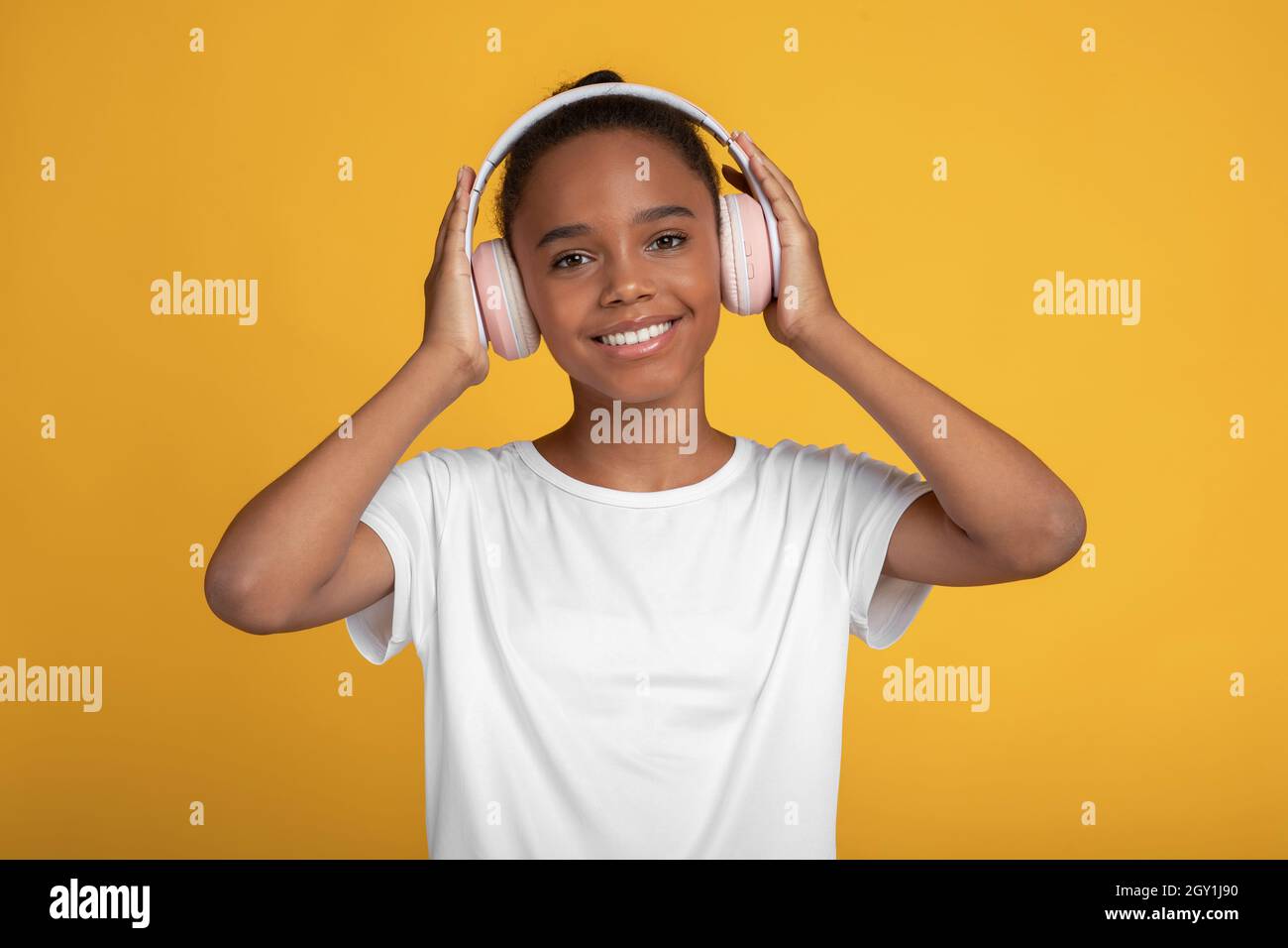 Allegra bella ragazza afroamericana in t-shirt bianca in auricolari ascoltare musica, isolato su sfondo giallo Foto Stock