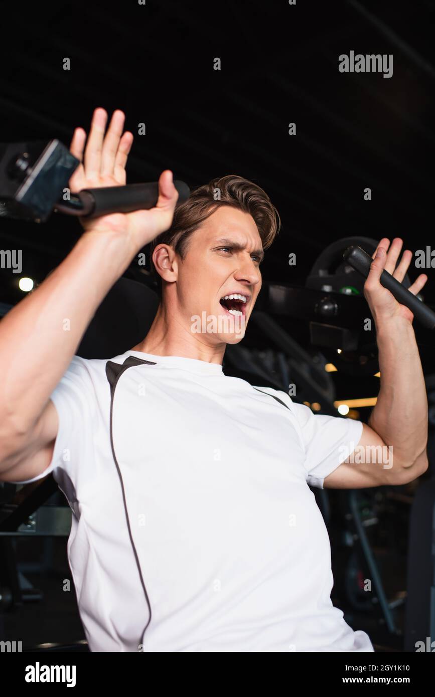 uomo sportivo che urla mentre si allenano sulla prolunga del braccio, esercitando la macchina nel centro sportivo Foto Stock