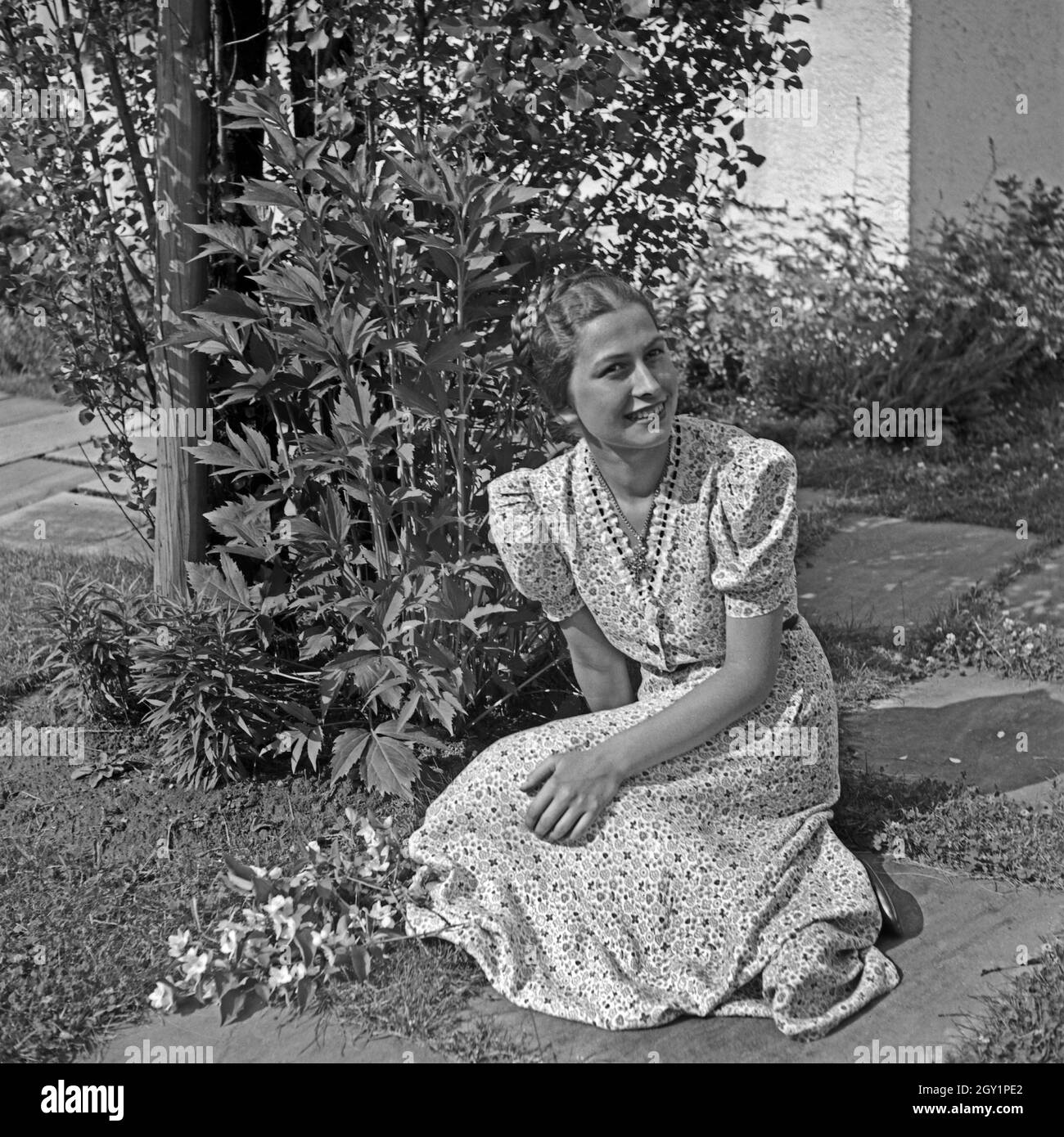 Eine junge Frau sitzt in einem Sommerkleid in ihrem Garten, Deutschland 1930er Jahre. Una giovane donna che indossa un vestito estivo è seduto nel suo giardino, Germania 1930s. Foto Stock
