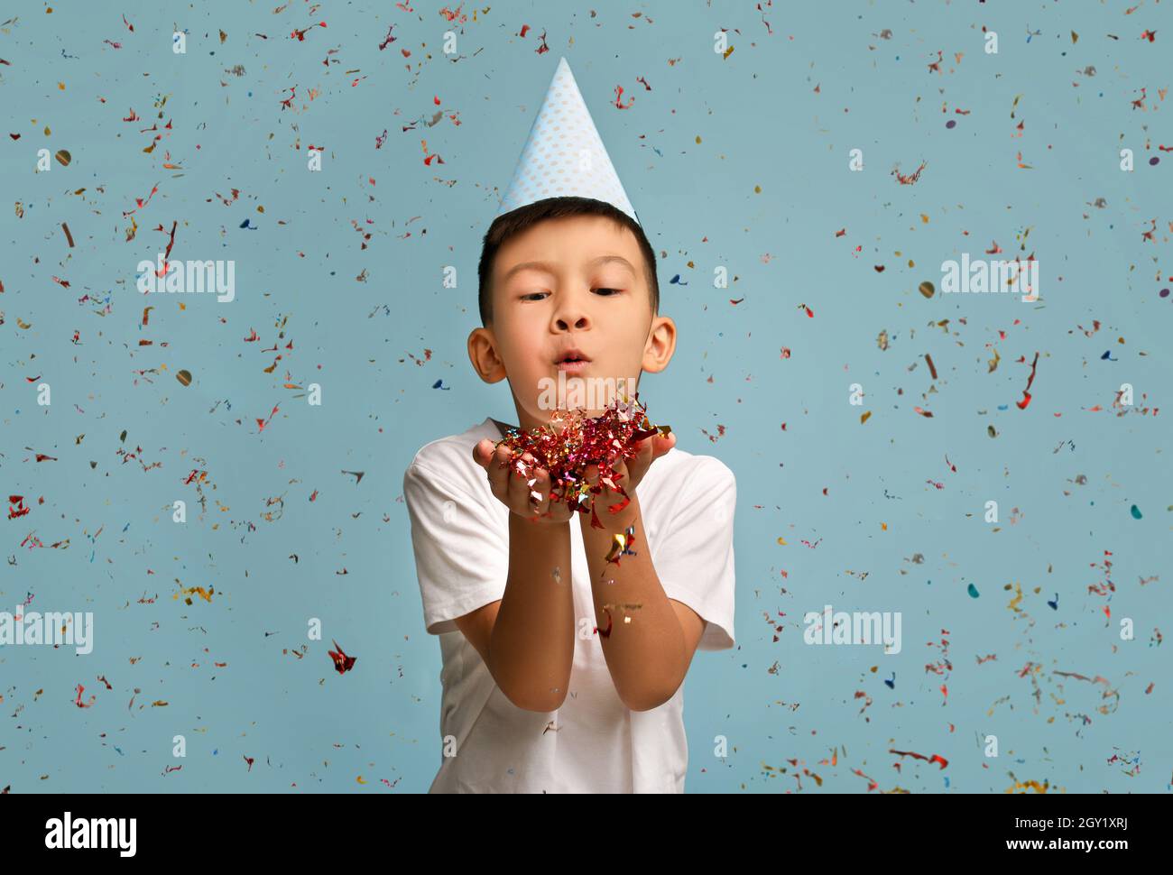 Buon compleanno. Little Asian Boy indossa il cappello di B-Day Blowing Confetti alla Camera Foto Stock