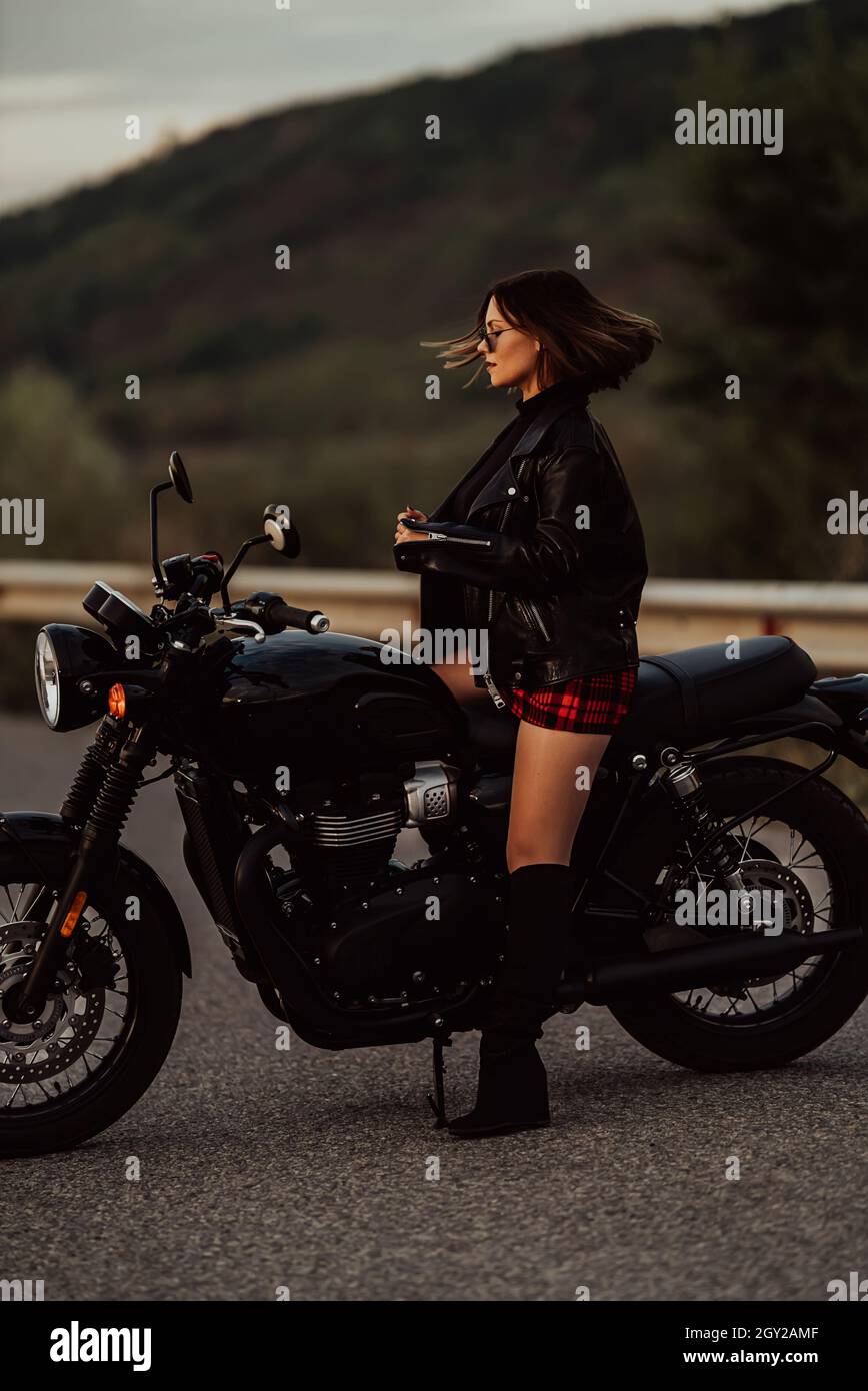 Ritratto di donna motociclista sicura in mini gonna, giacca in pelle si siede su una moto in stile retrò. Driver attraente in jackboot su autostrada Foto Stock