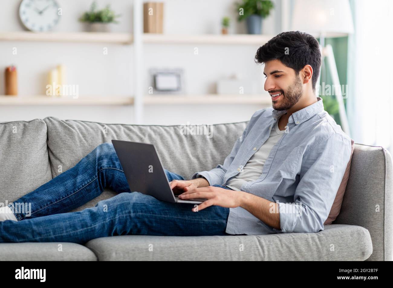 Felice ragazzo arabo che riposa con il laptop sul divano a casa, guardando film o i social network a scorrimento Foto Stock