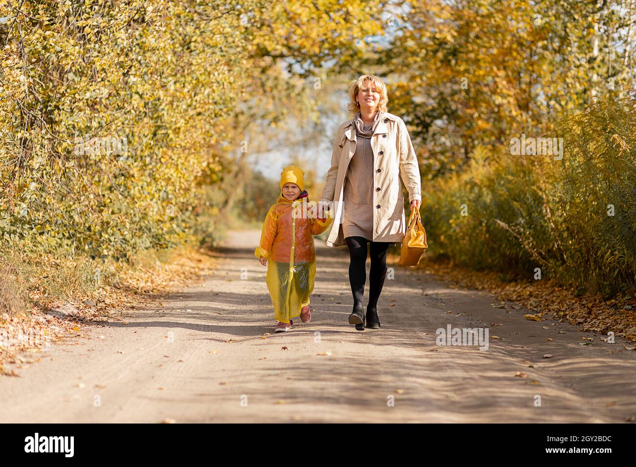 Foto di giovane donna cammina con bambina, figlia, 5 anni di tenuta mano su strada tra gli alberi, foglie gialle autunno Foto Stock