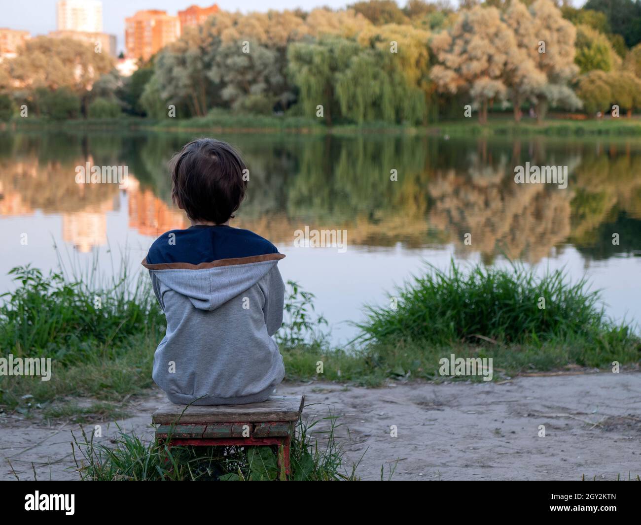 Il ragazzino è seduto a riva del lago e contempla. Persona seduta su una panchina da sola e guardando il tramonto Foto Stock