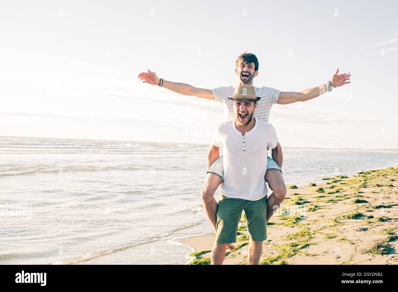 Due amici ragazzi ridono e sorridono e si divertono in spiaggia - ragazzo gioioso piggybacking su ragazzo giovane Foto Stock