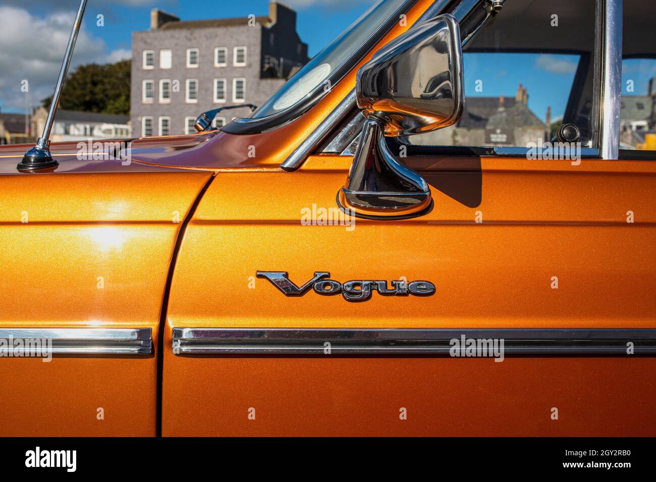 Vista laterale della porta passeggero e logo cromato su un'auto Sunbeam Vogue arancione metallizzata degli anni '70 sullo sfondo di edifici e cielo blu Foto Stock