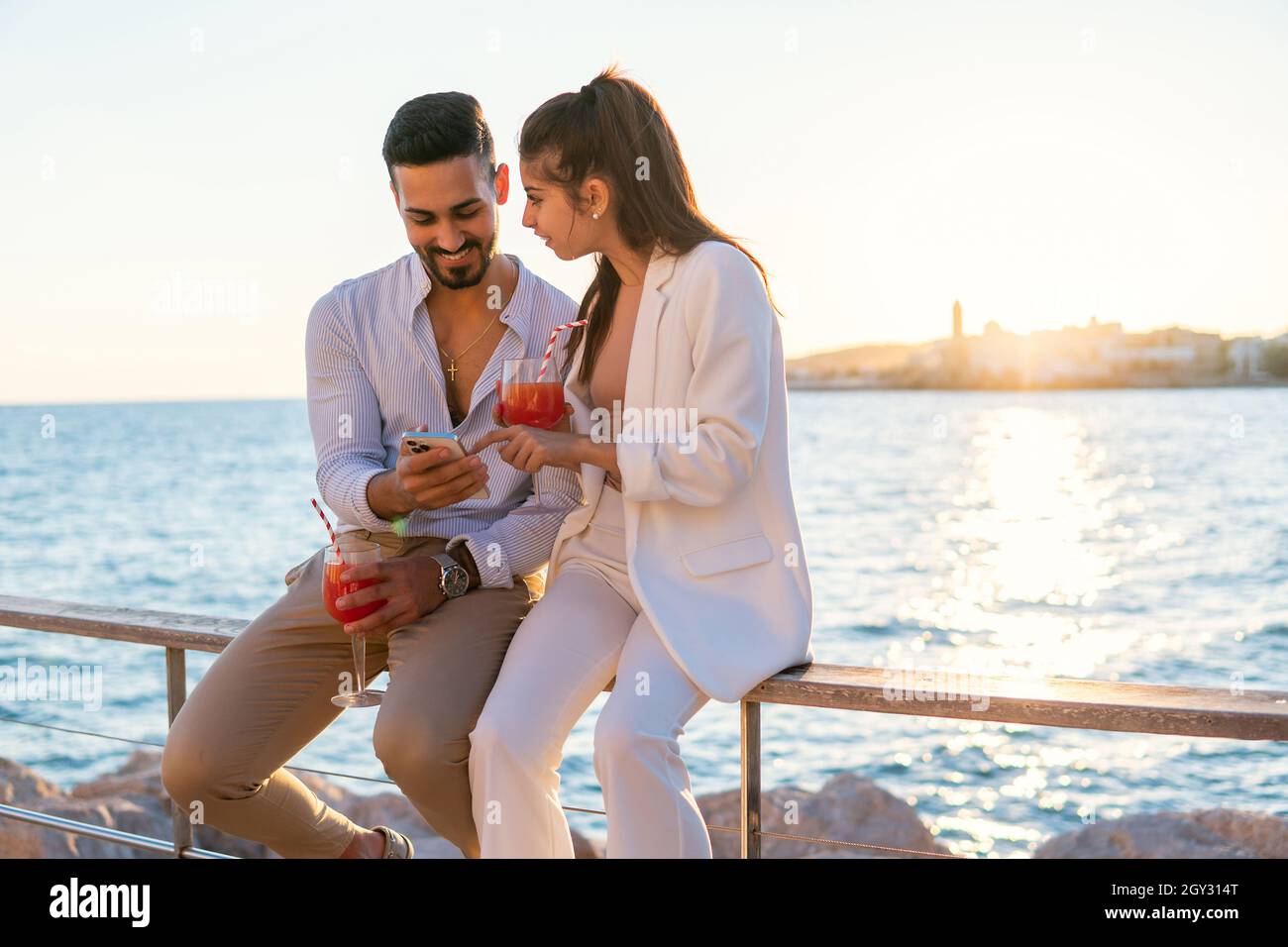 Una coppia etnico entusiasta con cocktail rinfrescanti in calici seduti sul lungomare e sullo smartphone mentre vi godete la serata estiva Foto Stock