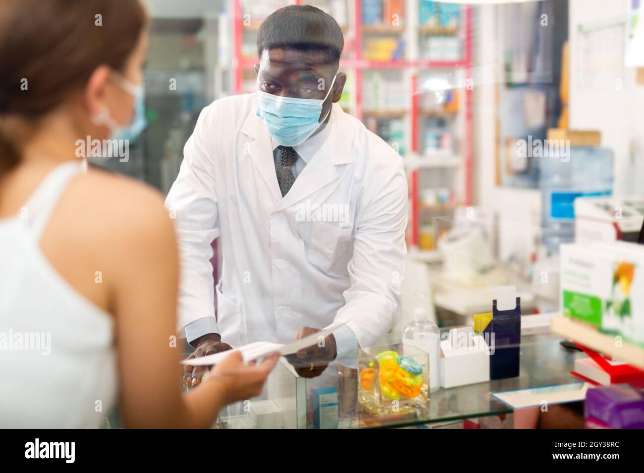 Il farmacista africano americano maschio in una maschera protettiva riceve una prescrizione per i farmaci da un cliente Foto Stock