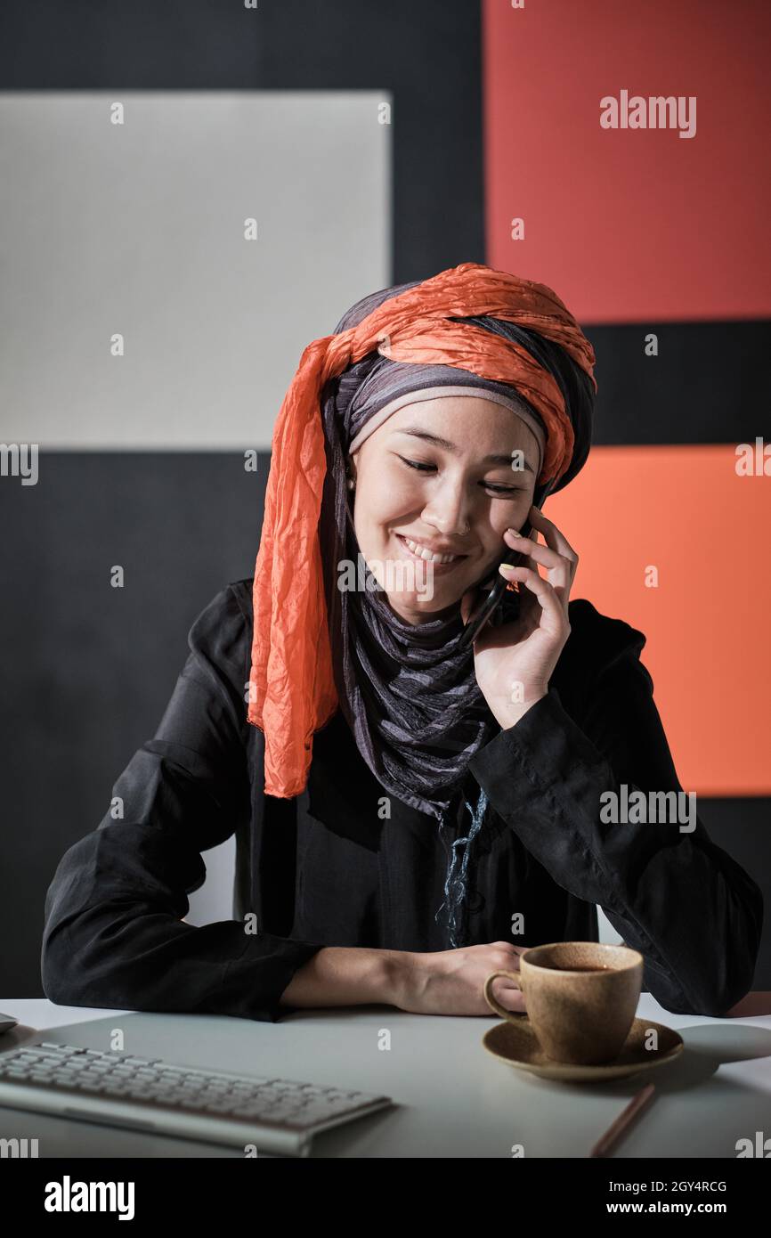 Donna d'affari di successo in abbigliamento musulmano seduto sul suo posto di lavoro in ufficio prendendo il telefono cellulare e sorridendo Foto Stock