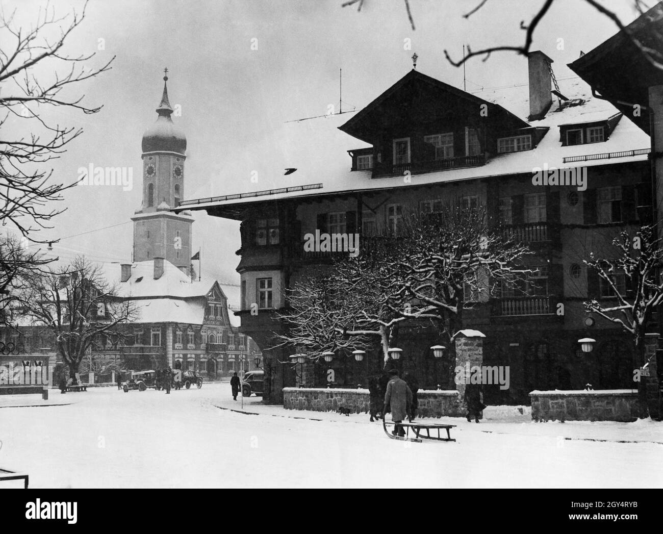 Il 4 febbraio, ha iniziato a nevicare a Garmisch-Partenkirchen. Proprio in tempo, perché la neve era necessaria per l'apertura delle Olimpiadi invernali il 6 febbraio e le seguenti competizioni. Nella foto, un uomo sta tirando la sua grande slitta attraverso la fresca neve nuova attraverso la Marienplatz, passato casa No. 13-15, che ha ospitato un hotel. Sulla sinistra dietro di esso la vecchia farmacia (casa no 10) e dietro di essa si può vedere la chiesa parrocchiale di San Martino. A sinistra, i poster pubblicitari per le Olimpiadi si appendono su un tabellone. [traduzione automatizzata] Foto Stock