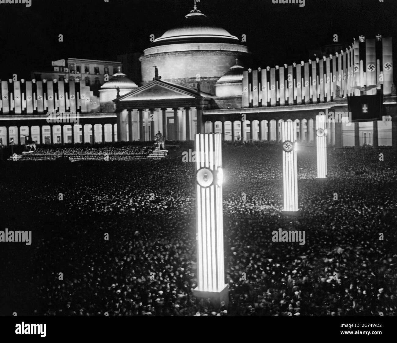 Una folla si è riunita nella luminosa Piazza del Plebiscito di Napoli la sera del 5 maggio 1938. L'occasione è stata Fleet Day in città. Sullo sfondo si trova la chiesa di San Francesco di Paola. Sulle colonne si trovano i seguenti simboli (dalla parte anteriore a quella posteriore): Stemma del Regno d'Italia, swastika, Fascis del Partito Nazionale fascista. [traduzione automatizzata] Foto Stock