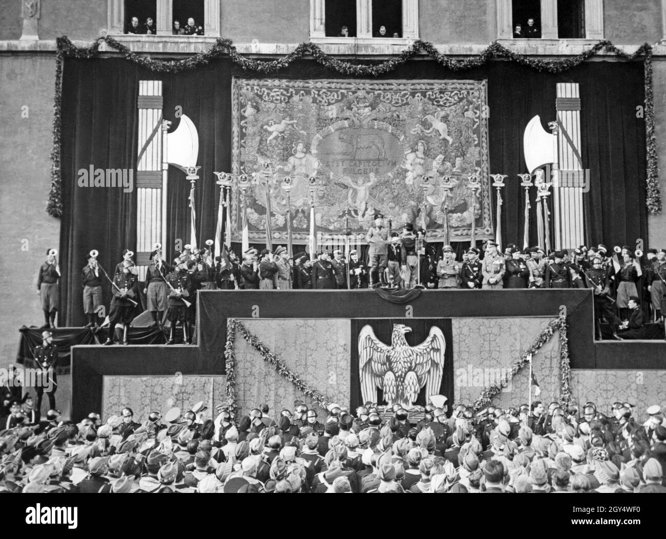 'Benito Mussolini (centro) ha tenuto un discorso ai suoi sostenitori in piedi in Piazza Venezia a Roma nel 1936. La facciata di Palazzo Venezia è drappeggiata da un telo ornato da due fasci di littori e da un arazzo raffigurante la lupa romana e l'iscrizione 'Stet capitolium fulgens' [raggiante il Campidoglio]. Alla destra di Mussolini ci sono tre rappresentanti del regime nazista tedesco. Prima del discorso Mussolini aveva premiato alcuni studenti che avevano combattuto nella guerra abissiniana. [traduzione automatizzata]' Foto Stock