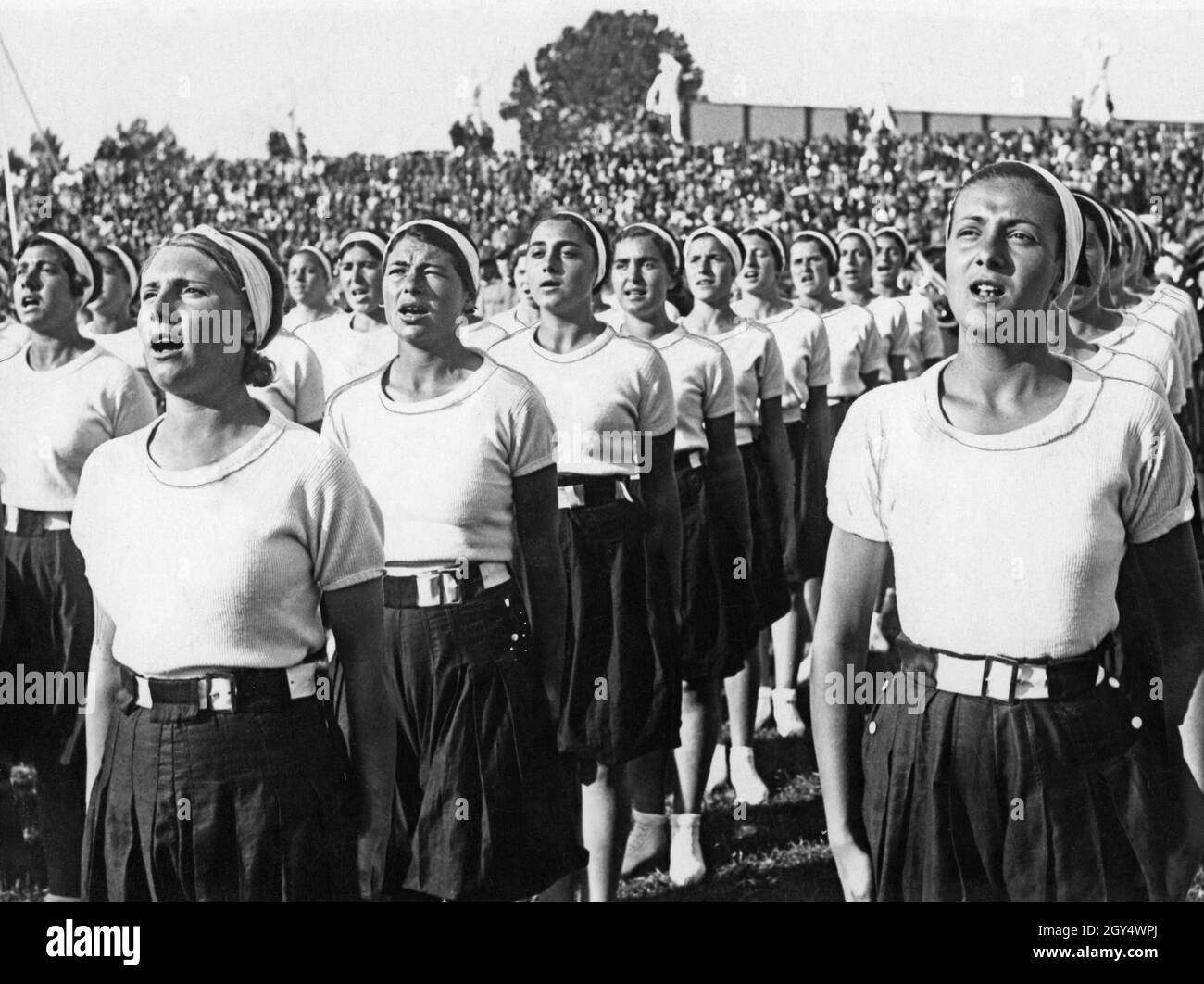Questa fotografia del 1933 mostra le giovani donne dell'organizzazione giovanile fascista Opera Nazionale Balilla cantando prima dell'inizio di un evento al Marble Stadium di Roma. Sullo sfondo si possono vedere delle bancarelle affollate. [traduzione automatizzata] Foto Stock
