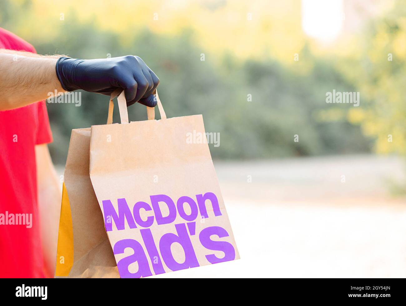 Una mano da uomo in guanto medico contiene una borsa di carta Mcdonal. Consegna a domicilio McCafe. Fast food McDonalds. Take away Mcdonal's. Foto Stock