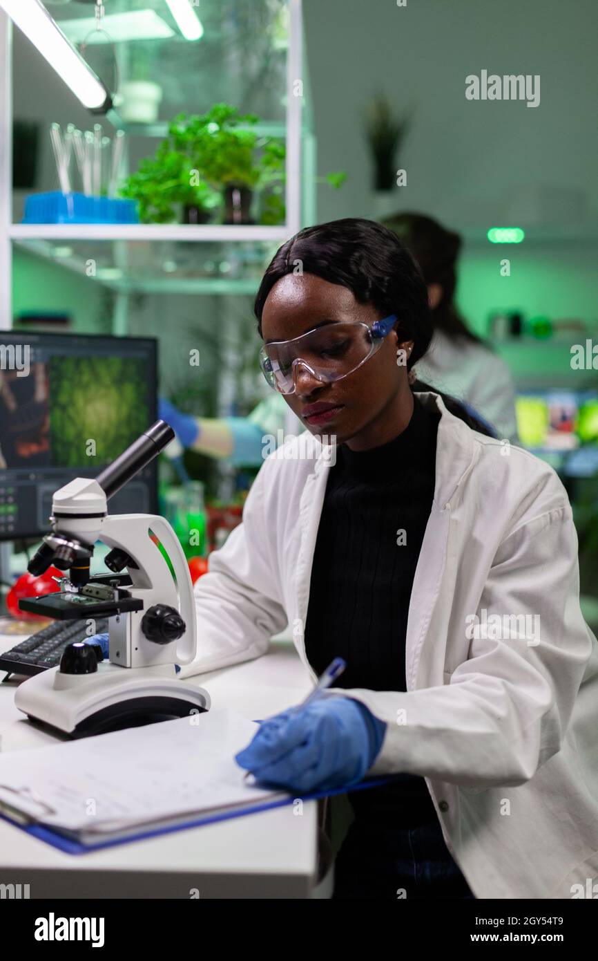 Il ricercatore botanico africano americano scrive i risultati dell'esperimento biologico sugli appunti dopo l'analisi della mutazione genetica su un campione vegetale al microscopio. Scienziato che lavora in laboratorio di biochimica Foto Stock