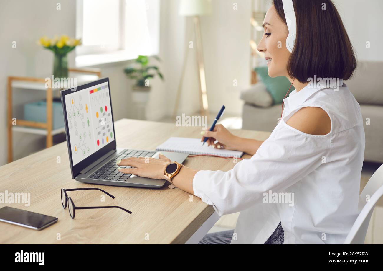 Donna seduta di fronte al portatile a casa, studiare online corso di business e prendere appunti Foto Stock