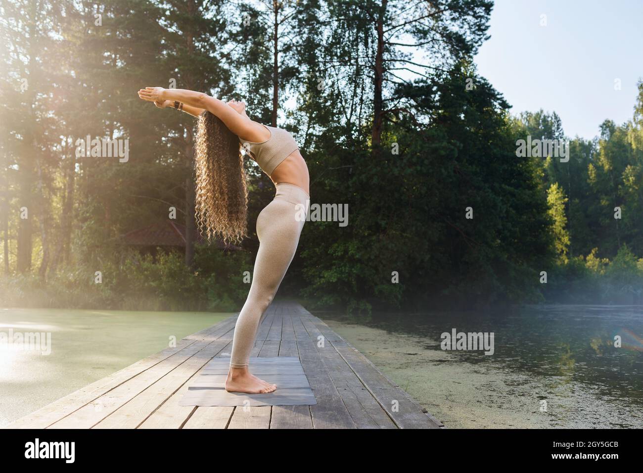 Una donna che pratica yoga svolge un esercizio, curva posteriore, posa Hasta Utttanasana, si erge su un ponte di legno in una calda mattinata di sole in un parco vicino a un pon Foto Stock