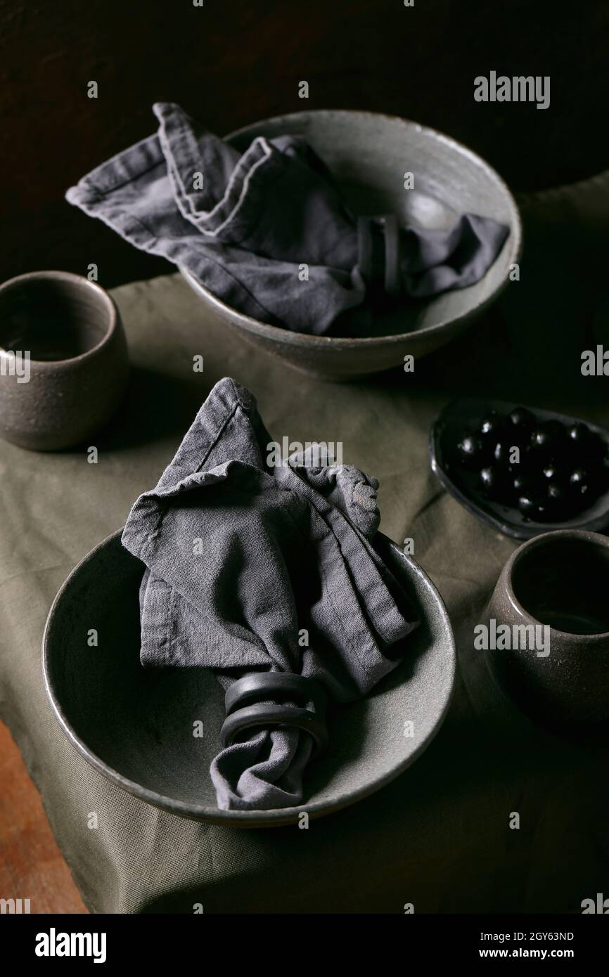 Ambiente da tavolo rustico con stoviglie in ceramica artigianale vuote, ciotole grezze grigie e tazze su tovaglia di lino. Sfondo scuro Foto Stock