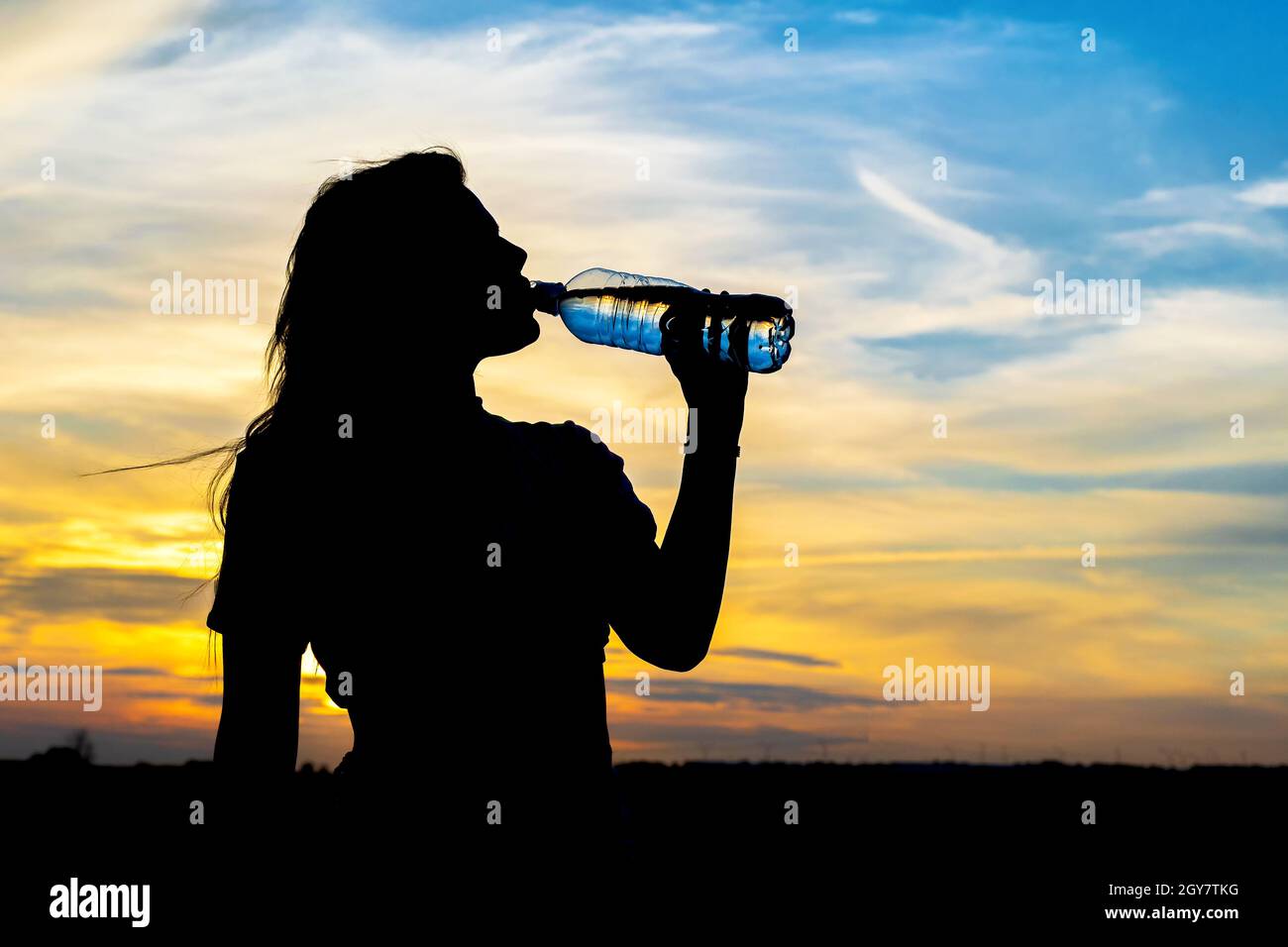 Silhouette nera di giovane donna sullo sfondo del cielo estivo del tramonto. Beve acqua dalla bottiglia. Foto Stock