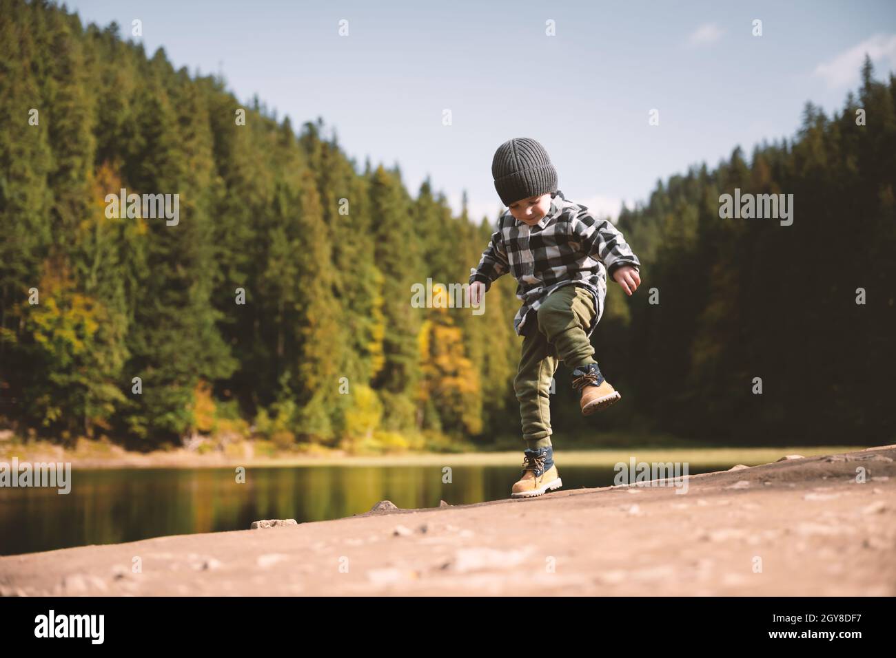Bambino piccolo in una camicia a quadri e cappello grigio sulla costa del lago forestale. Infanzia con natura concetto di amore Foto Stock