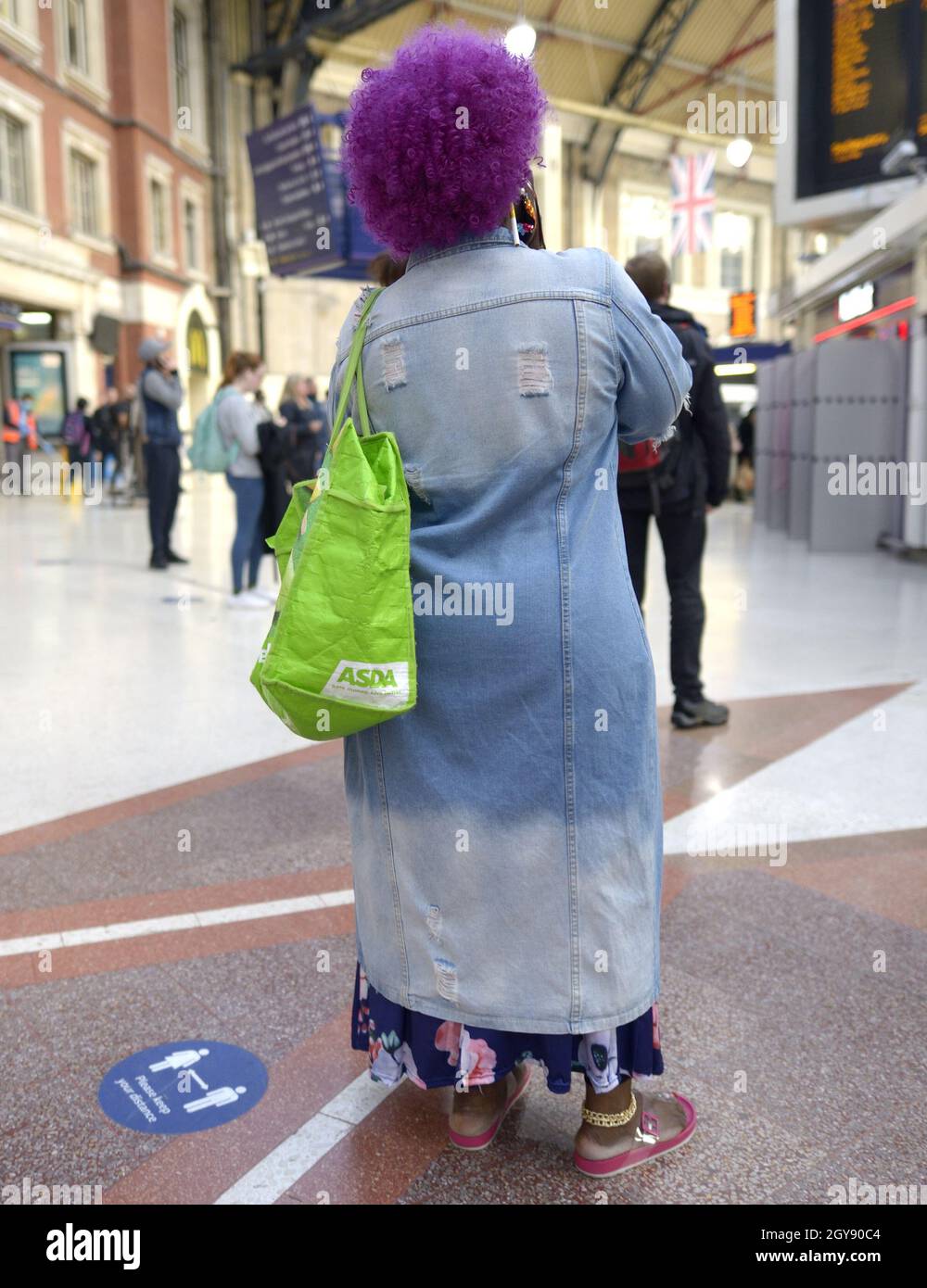 Londra, Inghilterra, Regno Unito. Victoria Station: Donna con capelli viola e borsa porta-porta Asda verde brillante che guarda il bordo delle partenze Foto Stock