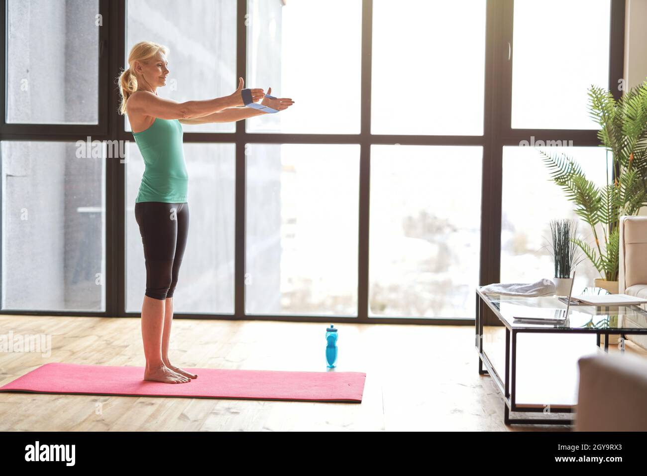 Vista laterale della donna bionda atletica in abbigliamento sportivo che si esercita utilizzando la banda di resistenza, seguendo il video tutorial online su un computer portatile, in piedi sul tappetino yoga Foto Stock
