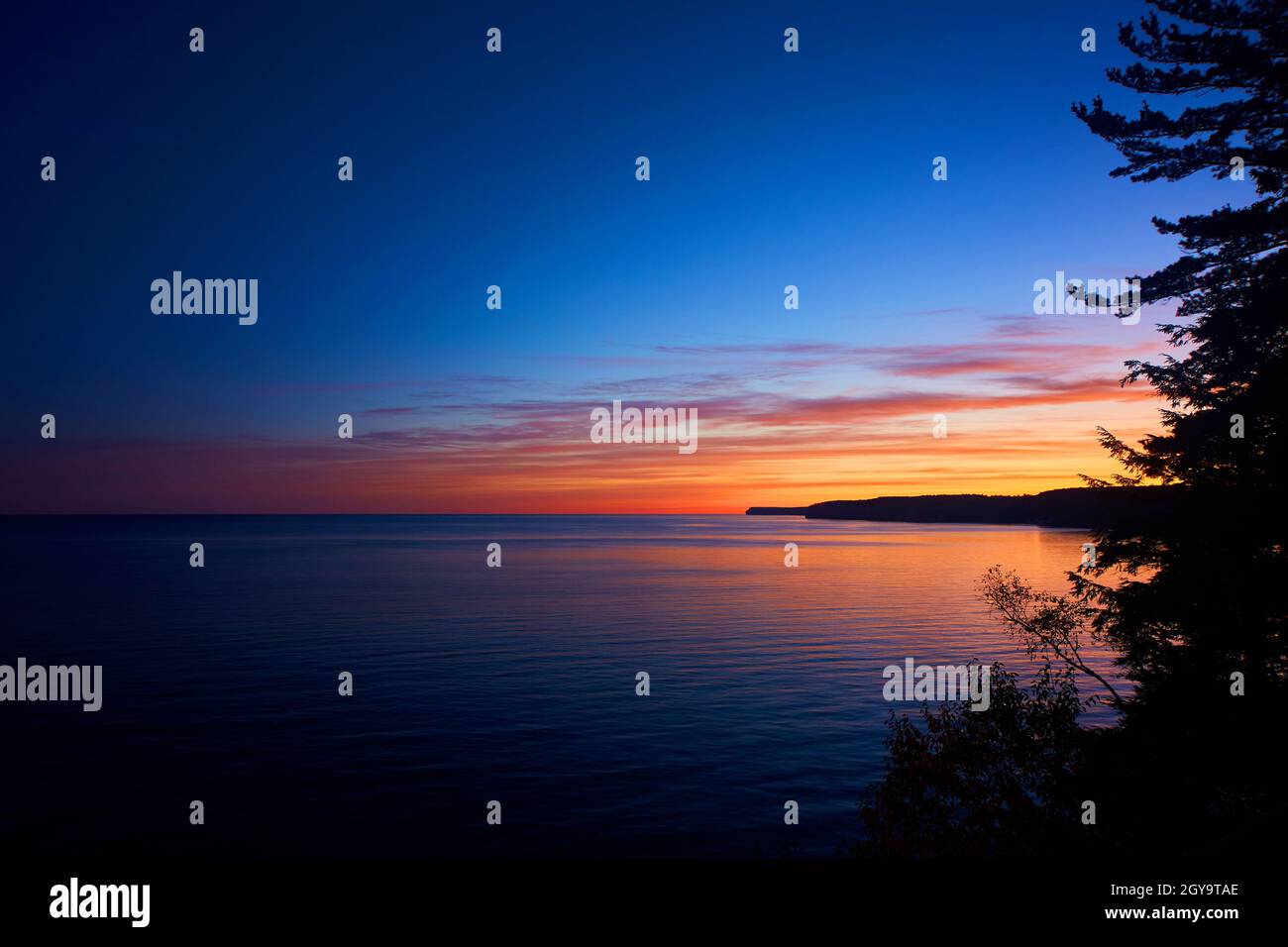 Acqua infinita illuminata da un'alba arancione rossa e gialla incorniciata su un lato dalla silhouette di un pino e di una penisola Foto Stock