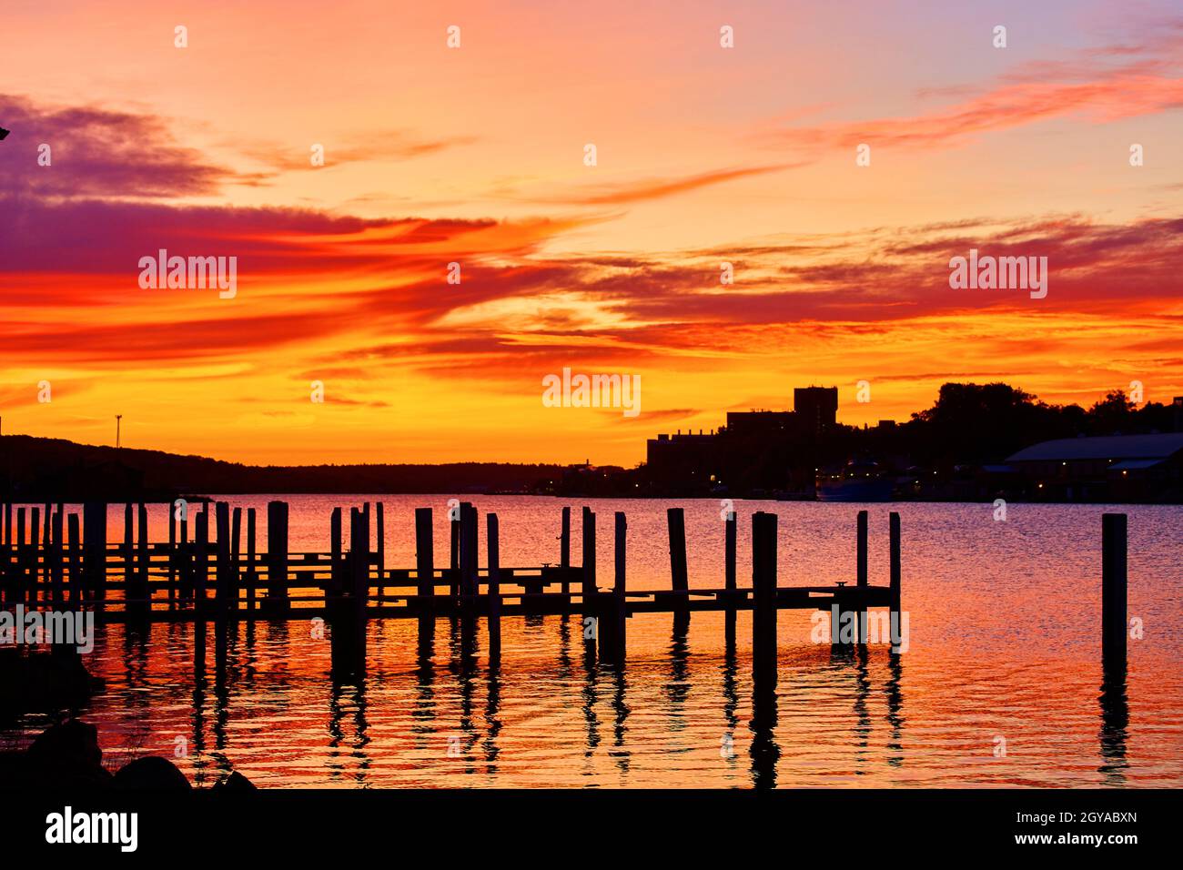 Brillante alba di rosso oro arancione e viola si riflette in un fiume d'acqua con sagome di banchine vuote e una città con luci Foto Stock