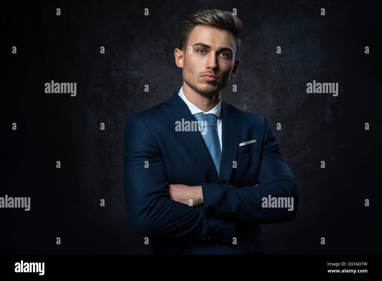 Ritratto concettuale di un giovane uomo d'affari di stile, in abito blu, mani su una croce, su uno sfondo scuro testurizzato. Foto Stock