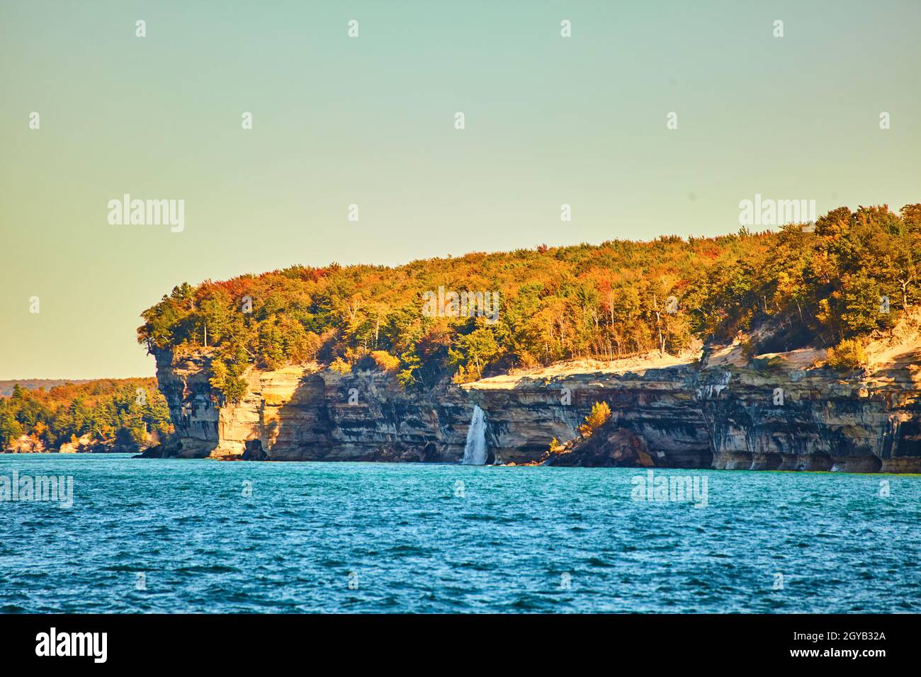 Rocce raffigurate con cascata distante che si riversa nel lago con alberi di caduta e onde ondeggianti Foto Stock
