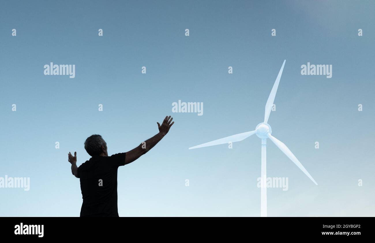 Uomo con le braccia aperte contro il cielo limpido guardando verso la turbina eolica. Foto Stock