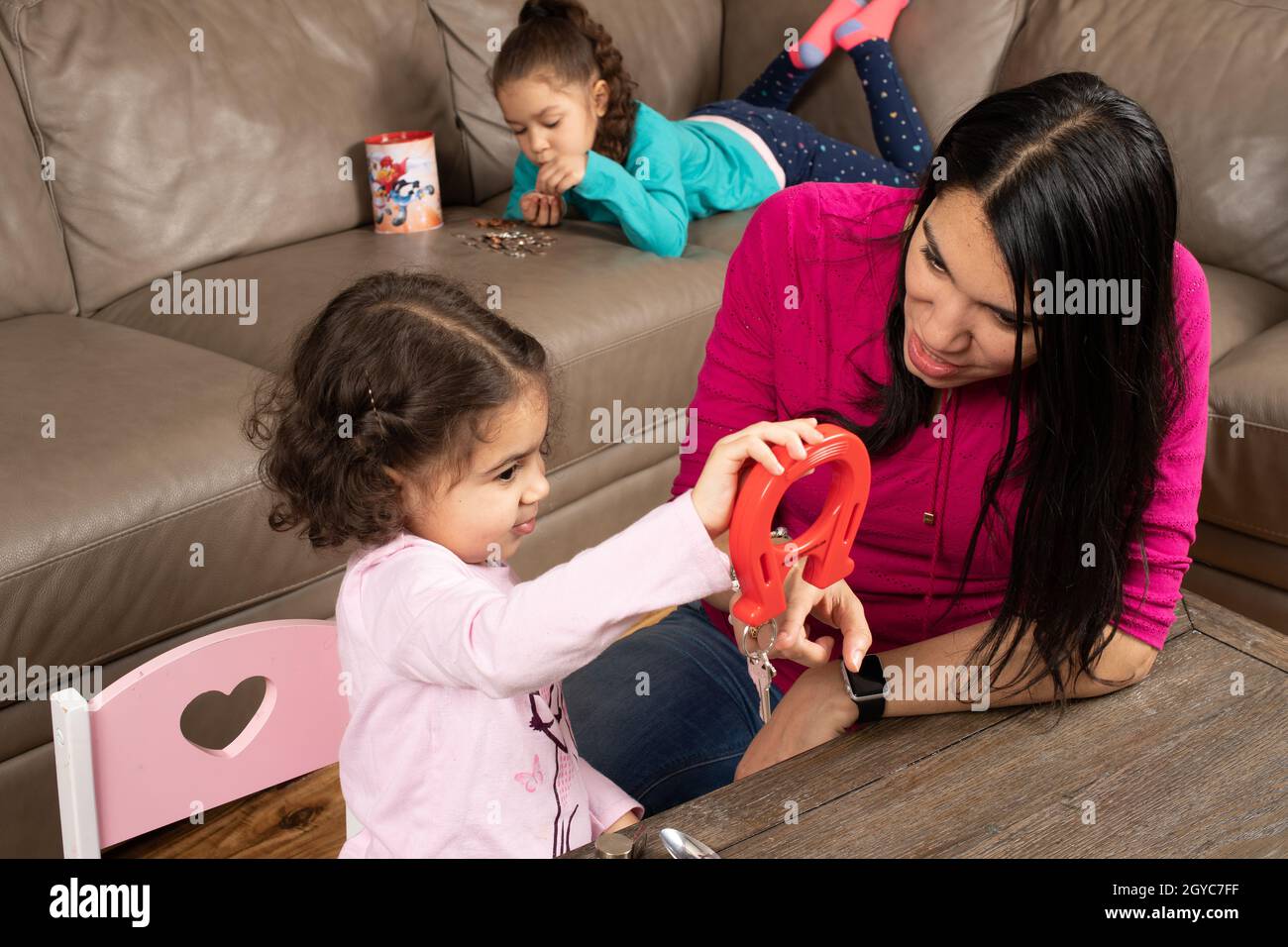 Madre incoraggiante figlia di 3 anni che gioca con magneti e oggetti metallici, come figlia di 5 anni conta e ordina le monete dalla sua banca di risparmio in b Foto Stock