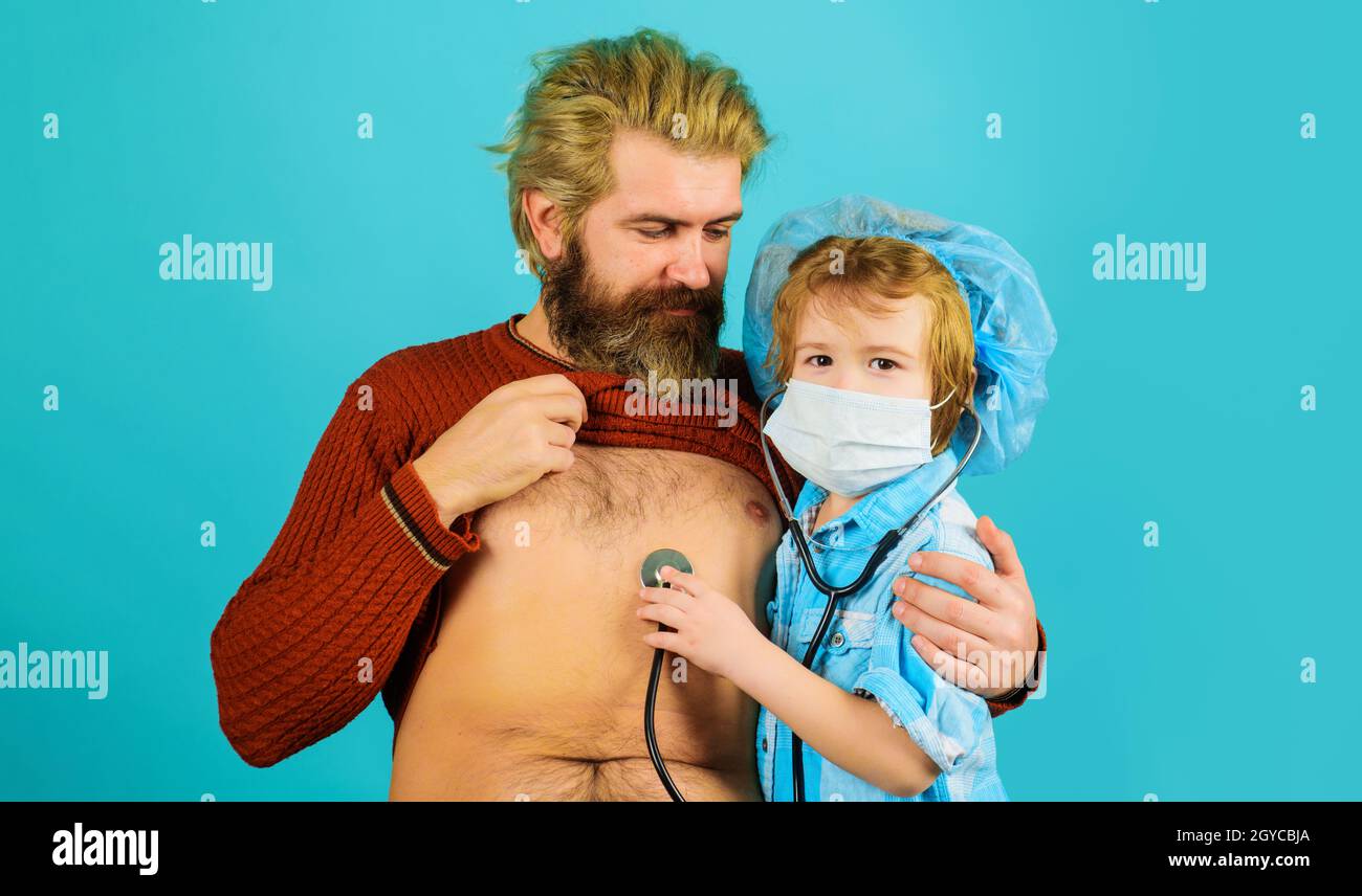 Famiglia che gioca in medico e paziente. Figlio in maschera medica con stetoscopio che ascolta il battito cardiaco del padre Foto Stock