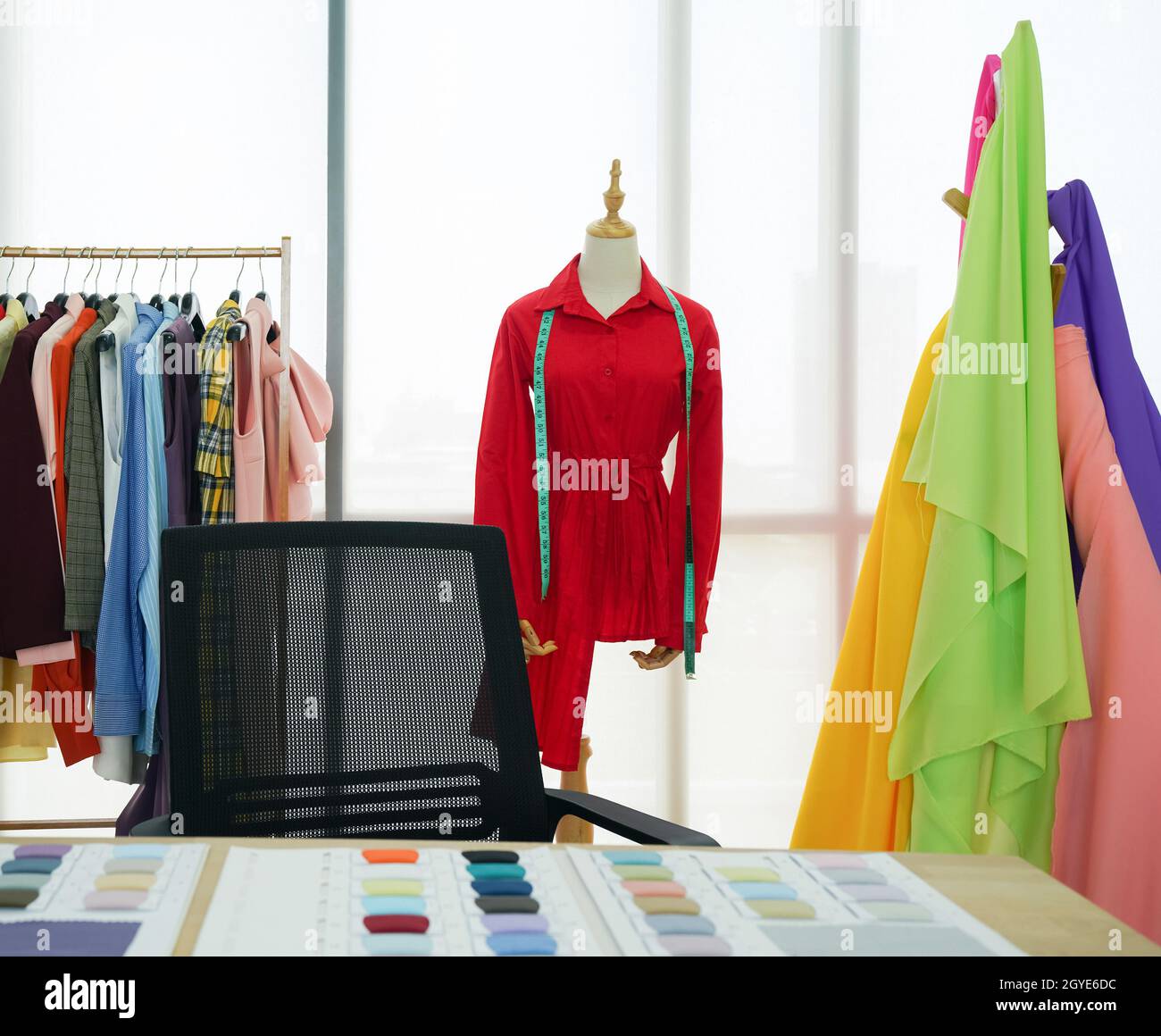 La stanza del designer di moda, la camicia rossa e il metro a nastro sono sul manichino. Un portabiti con tessuti colorati davanti alla grande finestra. Mattina Foto Stock