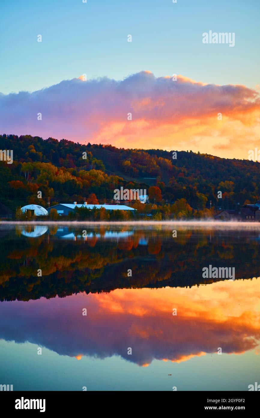 Verticale di un azzurro cielo alba con viola rosa e arancione nuvole con uno specchio di acqua e una casa o fienile con alberi di caduta Foto Stock