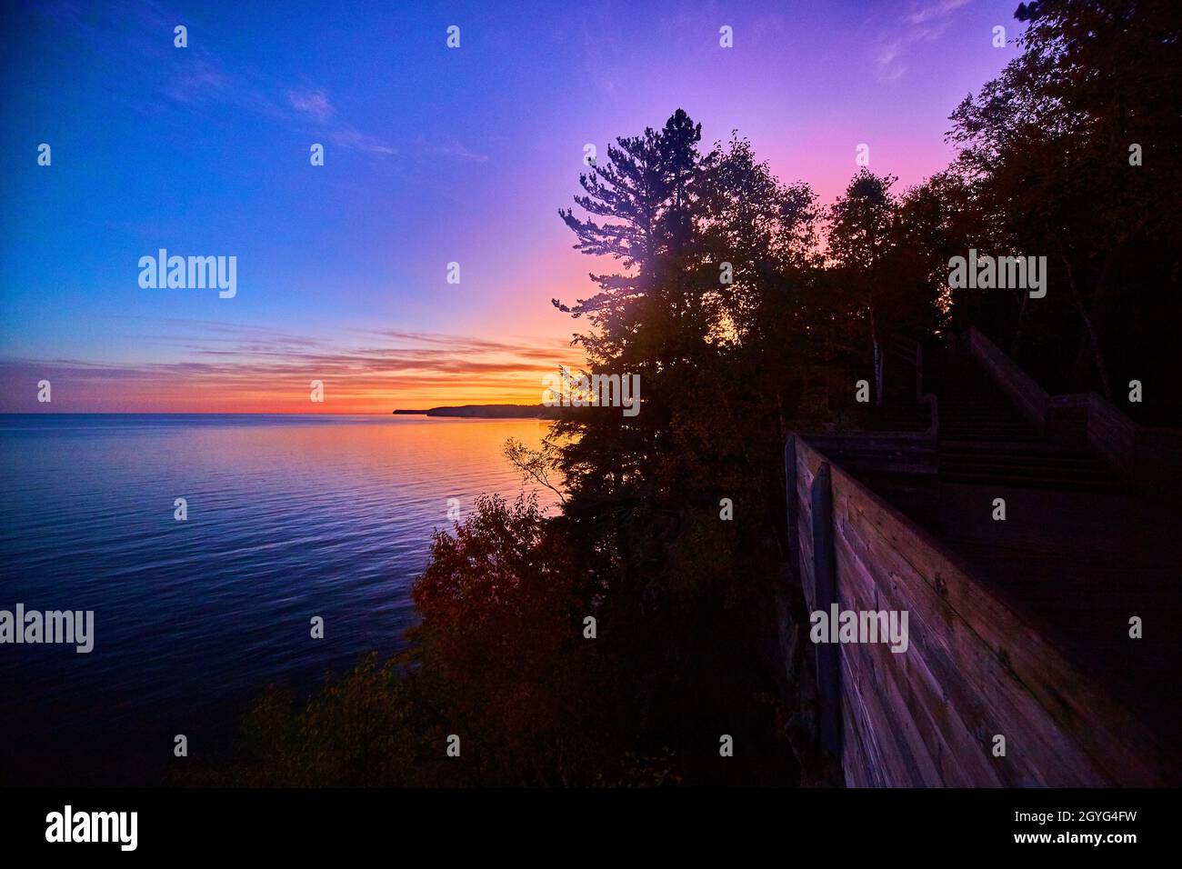 Alba d'oro, rosso, viola, rosa e blu su un lago o oceano con un sentiero di legno a destra che taglia attraverso una foresta Foto Stock
