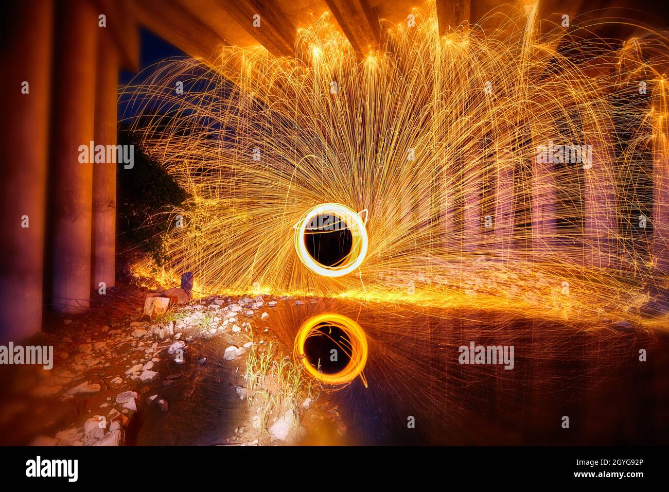 Portale di scintille gialle e bianche di luce da lana d'acciaio riflessa in acqua sotto un ponte di cemento Foto Stock