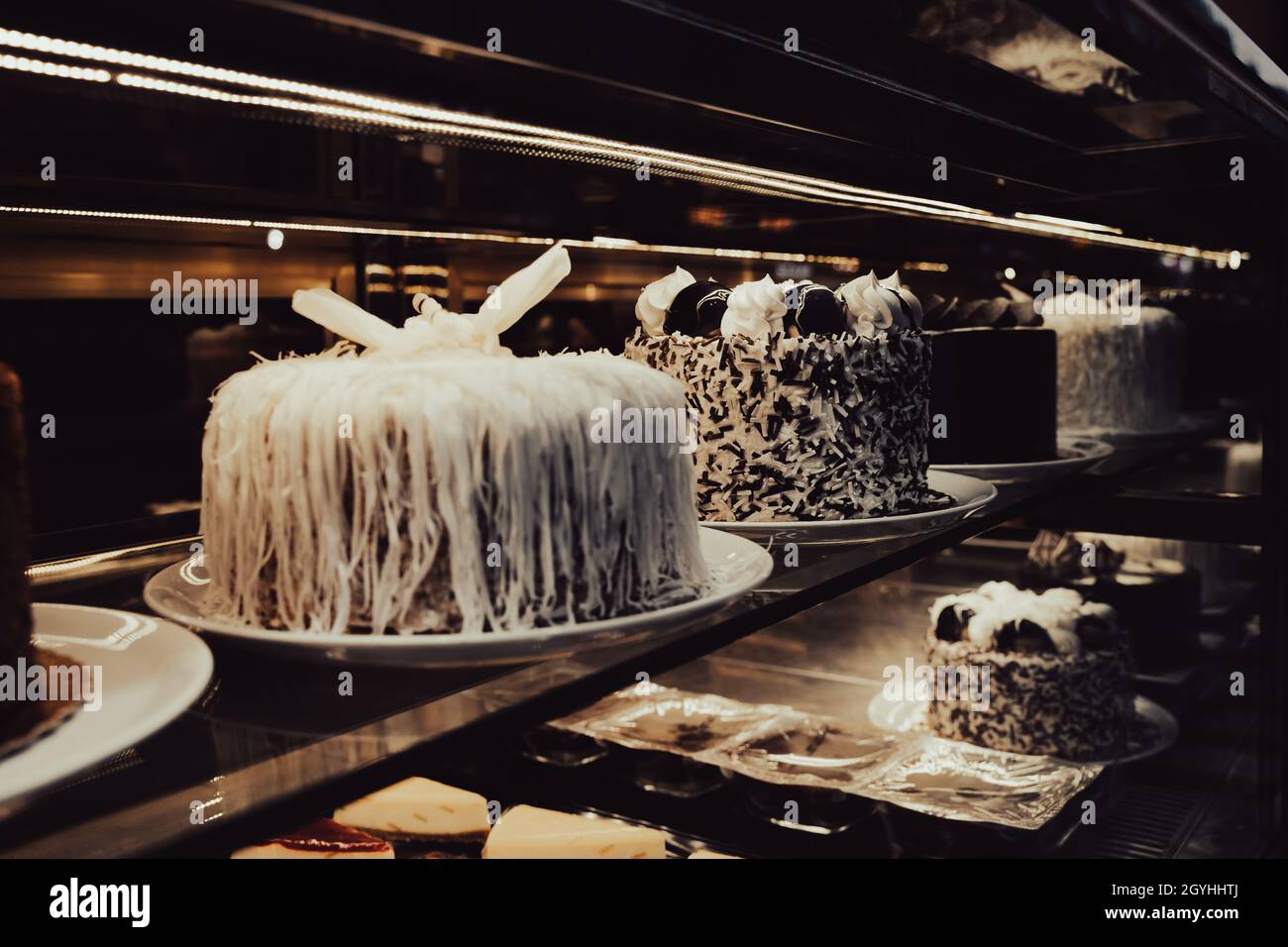 vista prospettica primo piano di file di torte di compleanno al cioccolato assortite allineate sullo scaffale nella finestra scura di un negozio di dolci Foto Stock