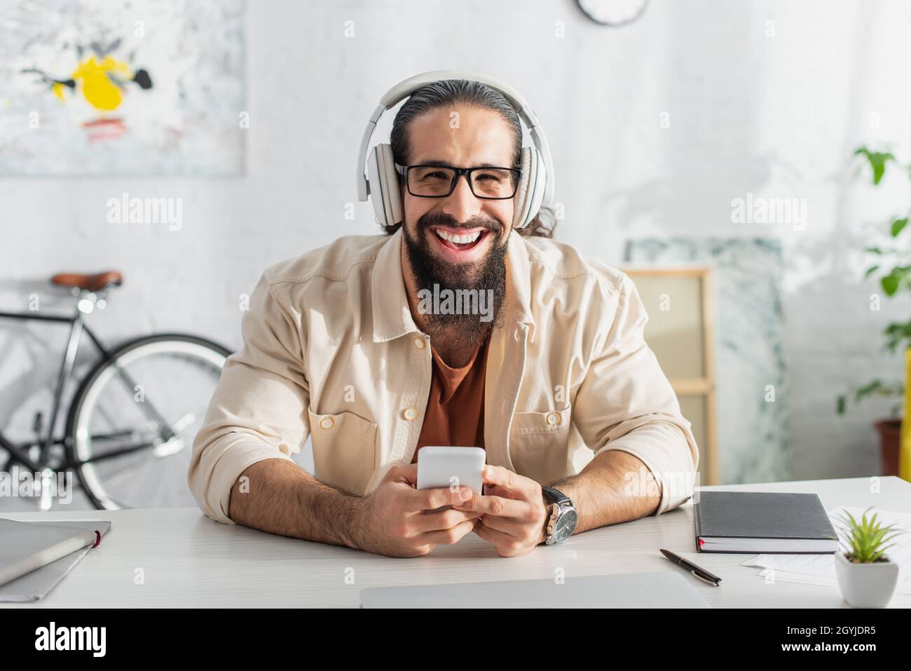 entusiasta freelancer ispanico in cuffie ridendo alla fotocamera mentre ascolti il podcast a casa Foto Stock