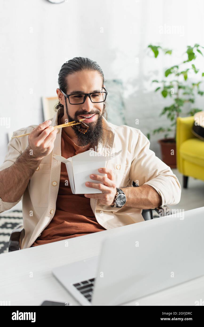 barbuto latino freelancer guardando il computer portatile mentre si mangia tagliatelle cinesi per pranzo a casa Foto Stock